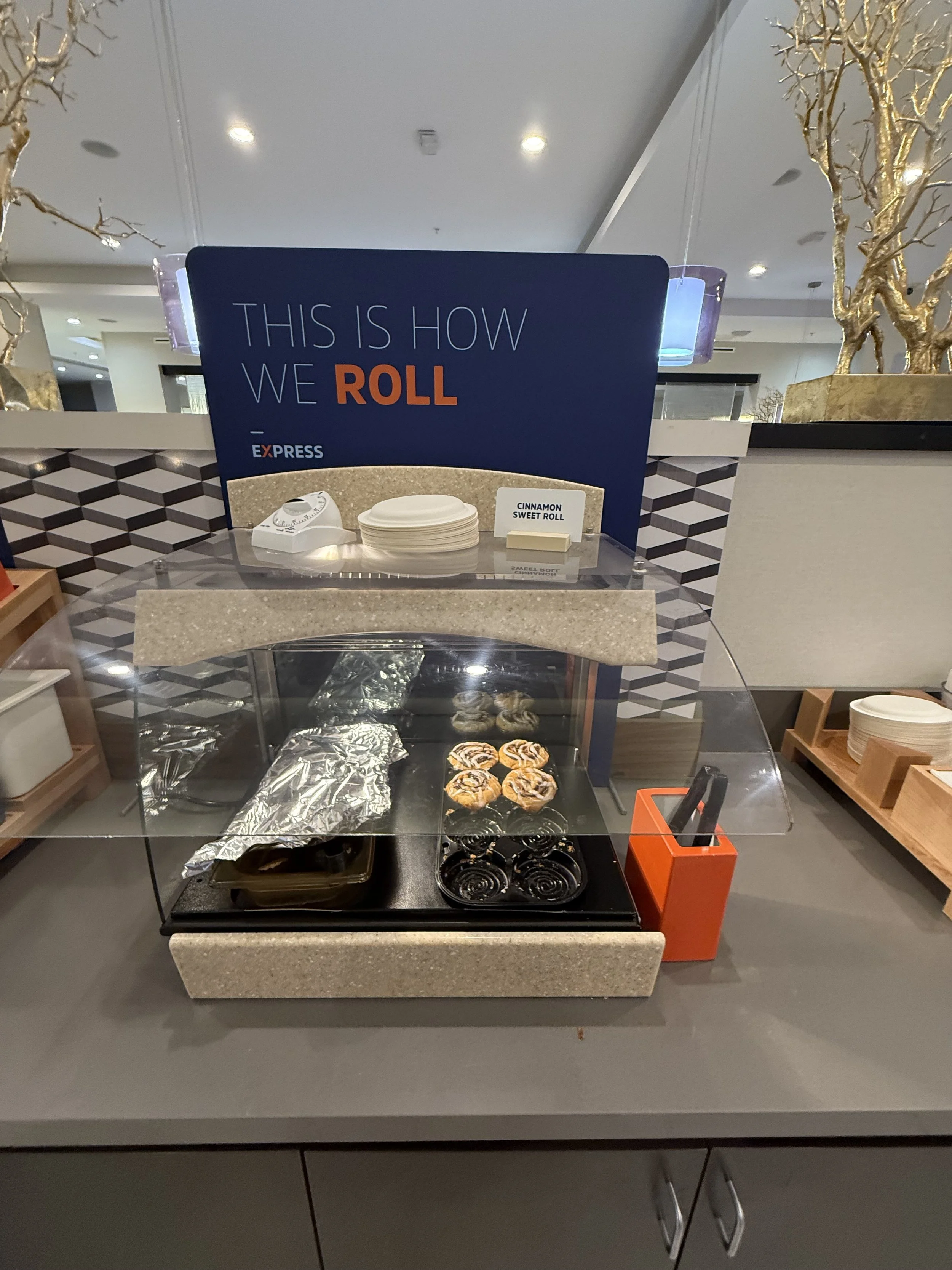 A cinnamon sweet roll display with baked rolls under a glass cover, a sign reading 'This is how we roll', and utensils in an orange holder, at an airport or food court.