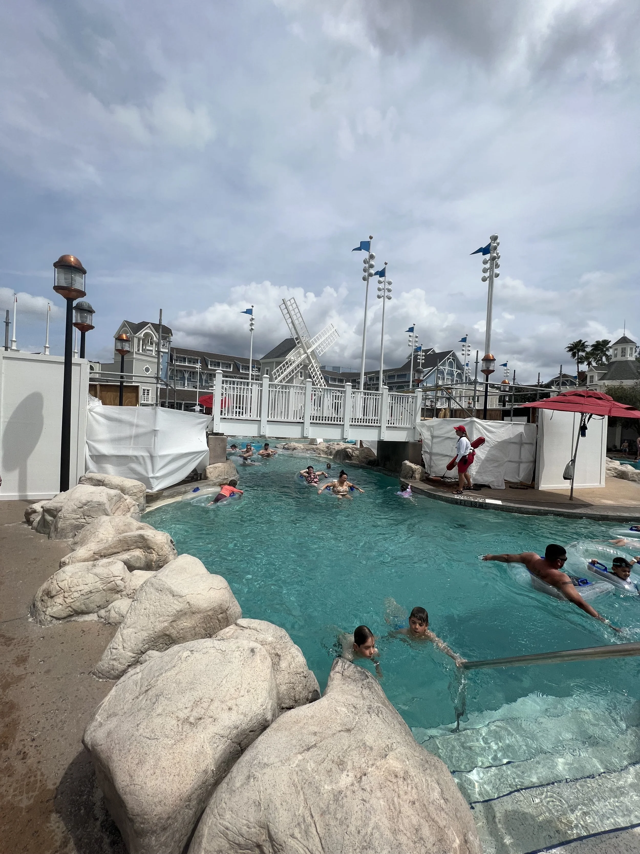 Children and adults swimming and playing in a large outdoor pool with rocks along the edge, bordered by white fencing and structures, under a cloudy sky, with a decorative windmill and buildings in the background.