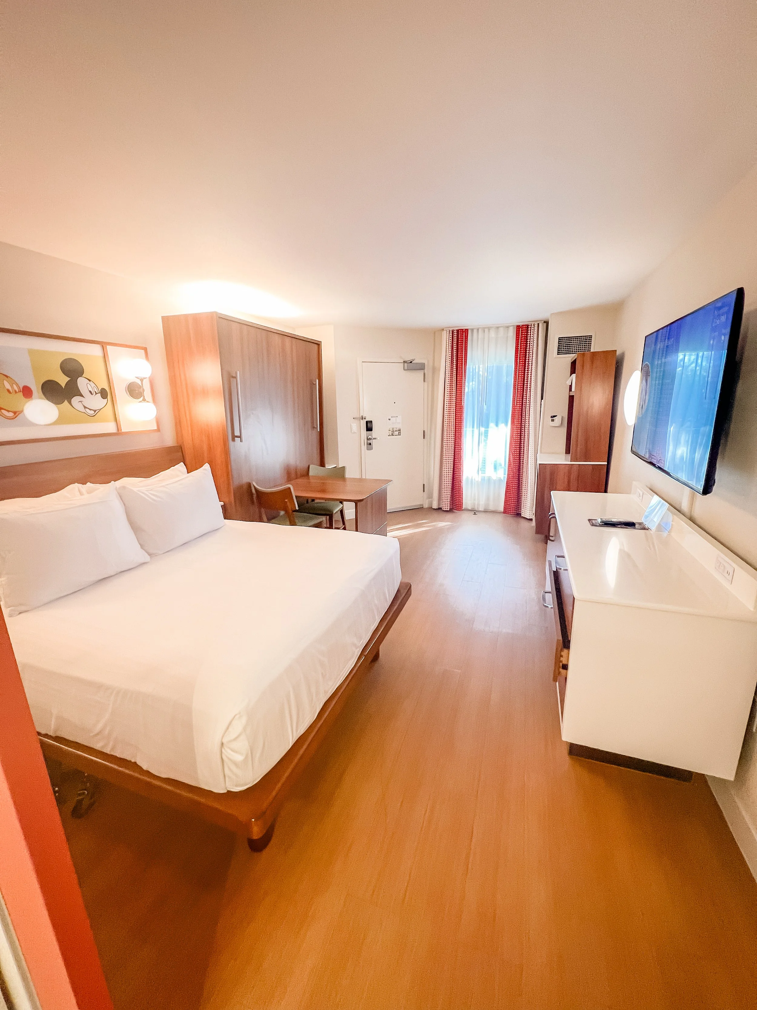 Hotel room with a large bed, wooden furniture, a wall-mounted TV, and a door with a window covered by curtains.