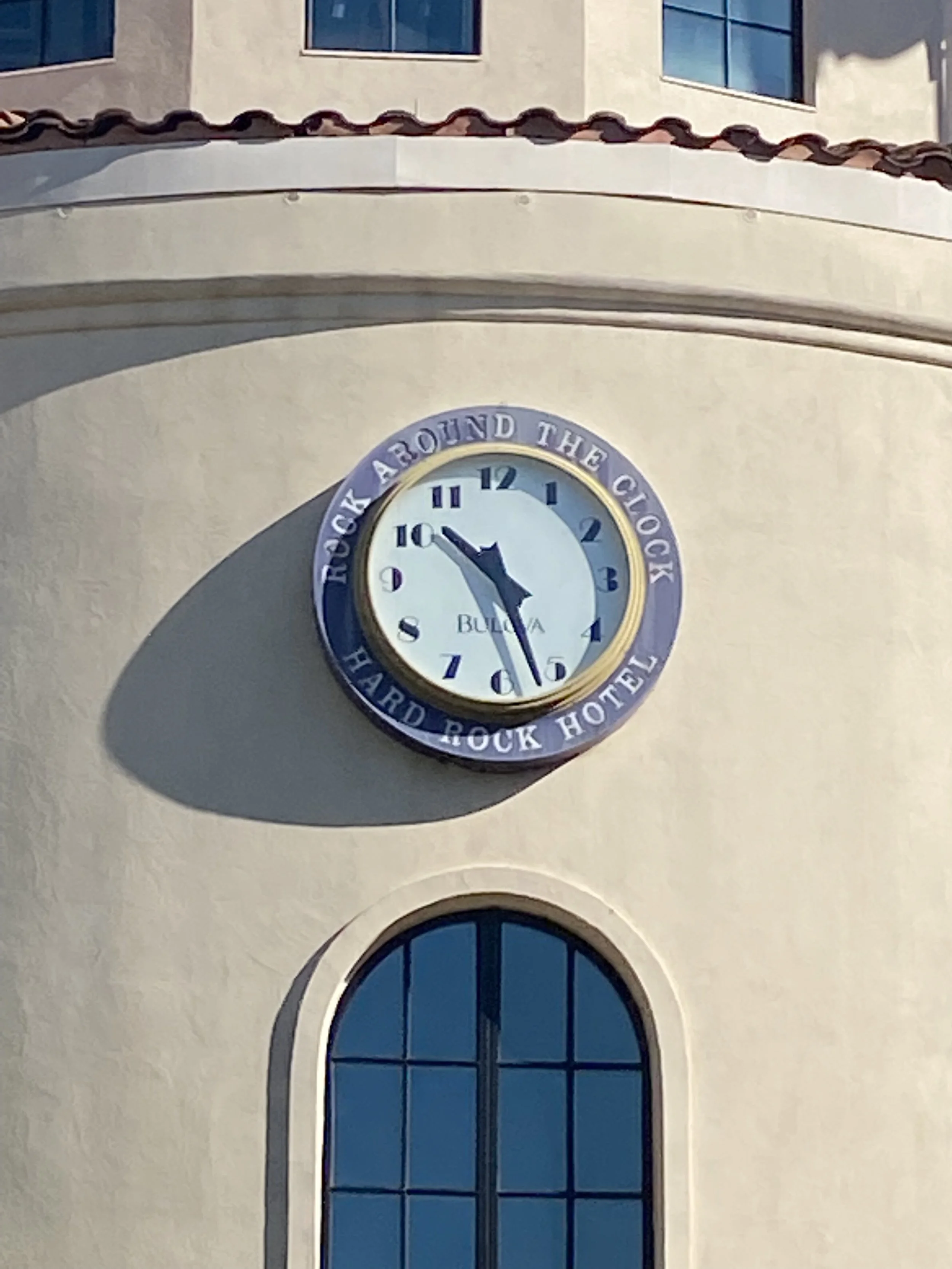 A clock on a building showing 4:50, with an outer ring that reads "Rock Around The Clock" and "Hard Rock Hotel." The building has a window below the clock and a roof with terracotta tiles.