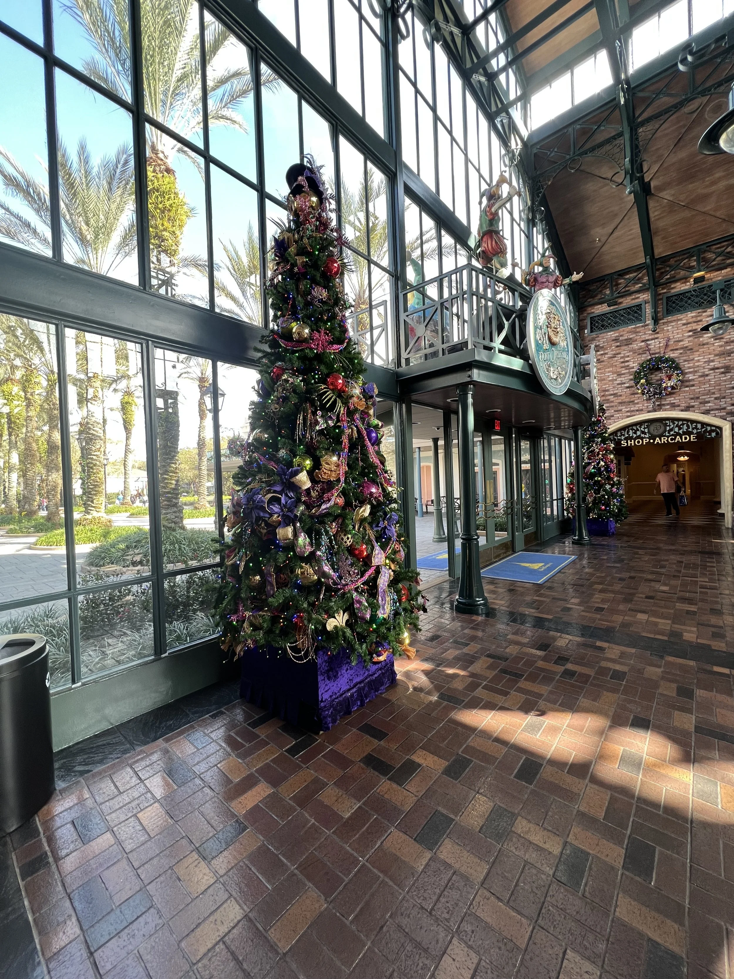 Decorated Christmas tree inside a glass-walled building, with palm trees visible outside. The interior has brick flooring and man-sized decor such as statues and signs, with a sign for an arcade shop in the background.