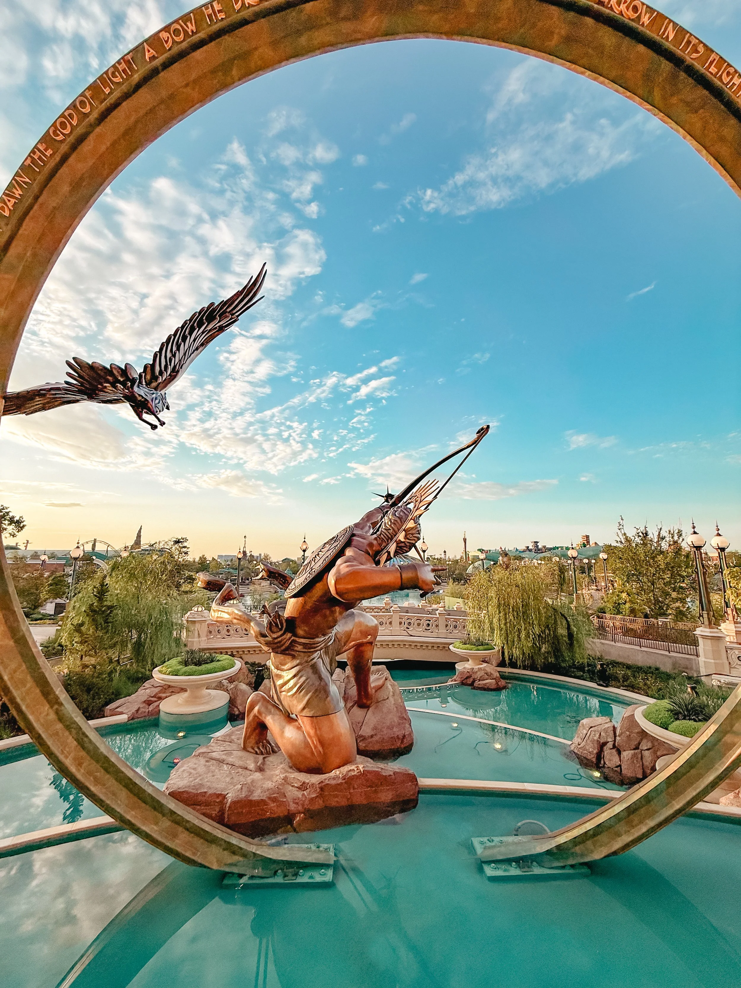 Bronze statues of a Native American man kneeling and shooting a bow, and a soaring eagle, framed by a large bronze circular arch with an inscription, with a scenic park and sky with clouds in the background.