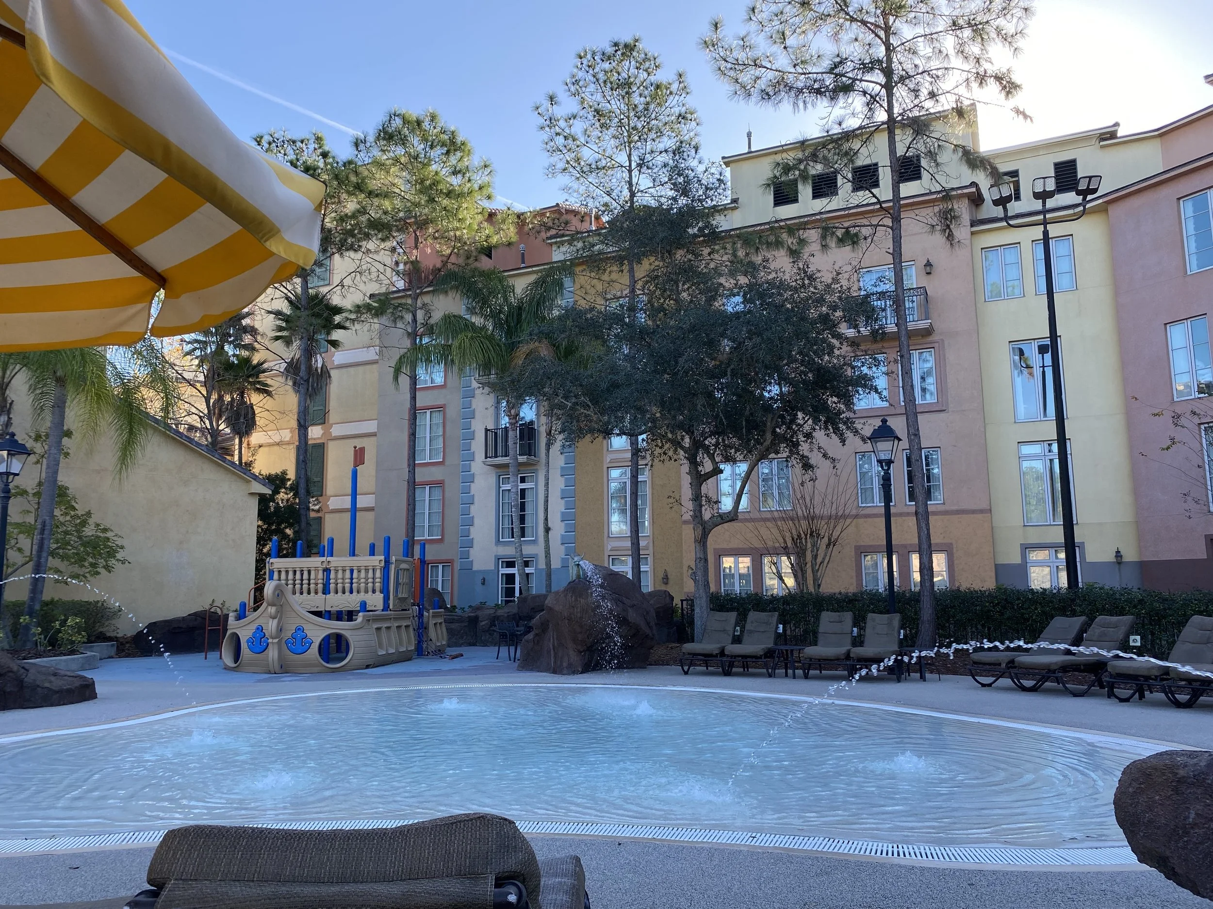 A swimming pool area with lounge chairs, large rocks, and a yellow and white striped umbrella, surrounded by a multi-story apartment building and tall trees, during daytime.
