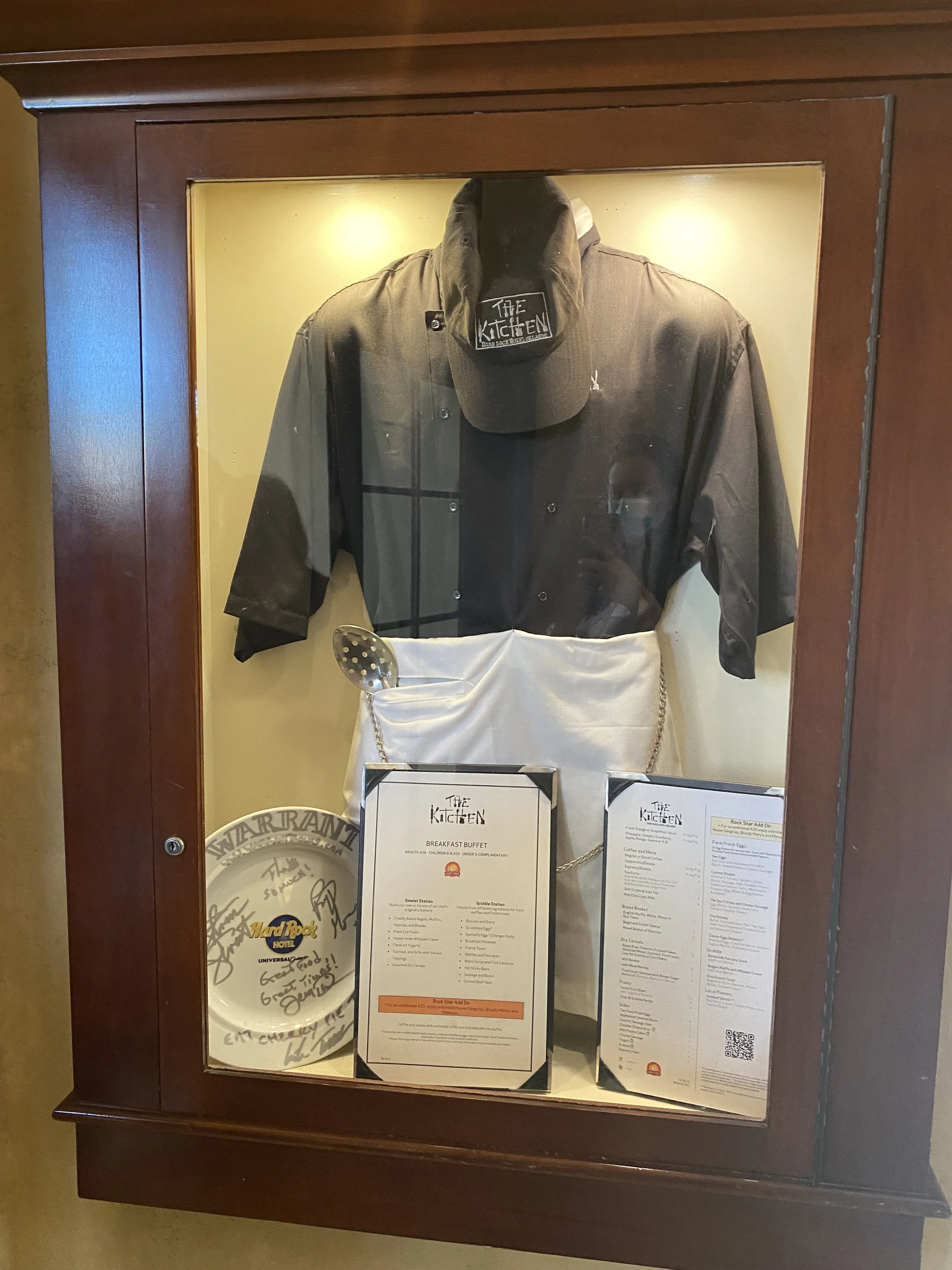 Display case with a chef's uniform, hat, and kitchen-related memorabilia, including signed items and menus from The Kitchen restaurant.