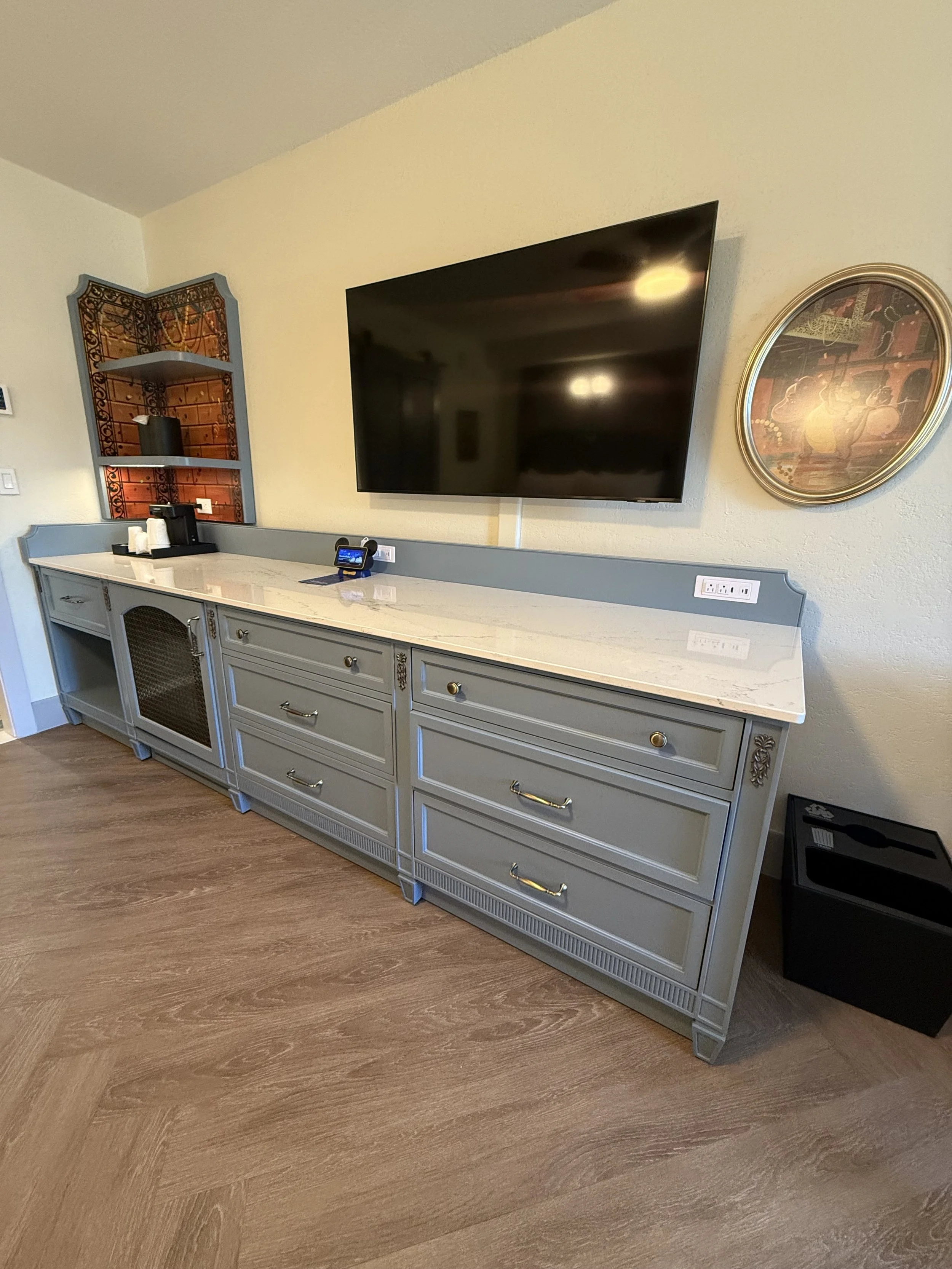 A sideboard with drawers and cabinets, topped with a marble surface, in a room with wooden flooring. A flat-screen TV is mounted on the wall above it. There are decorative items and a small device on the sideboard.