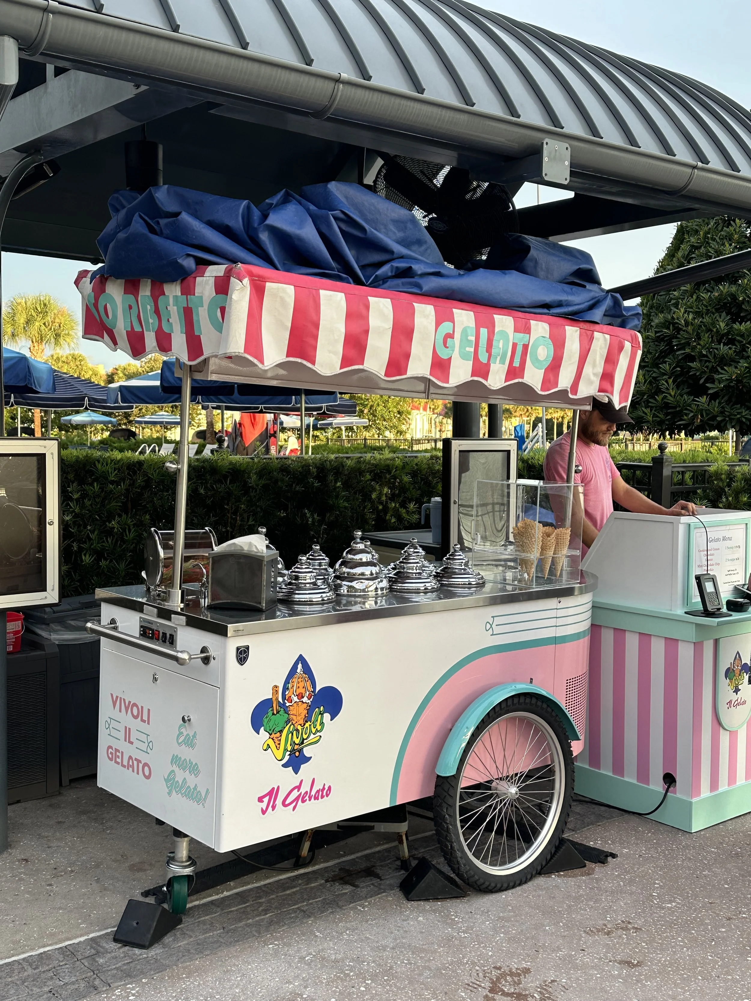 Ice cream cart with pink and white striped canopy selling gelato, with a man working behind the counter at an outdoor setting with trees and other carts in the background.