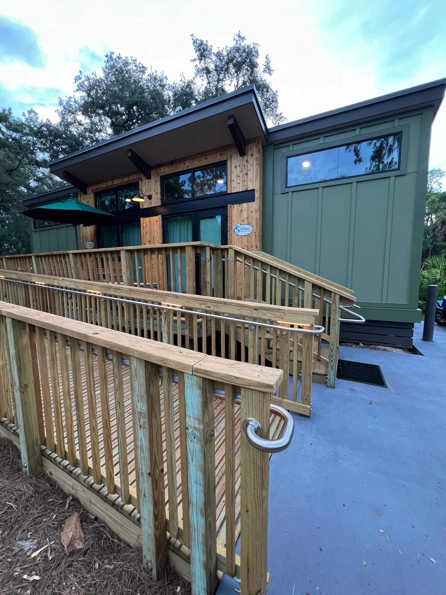 Exterior view of a contemporary building with a wooden ramp and railing at the entrance, surrounded by trees.
