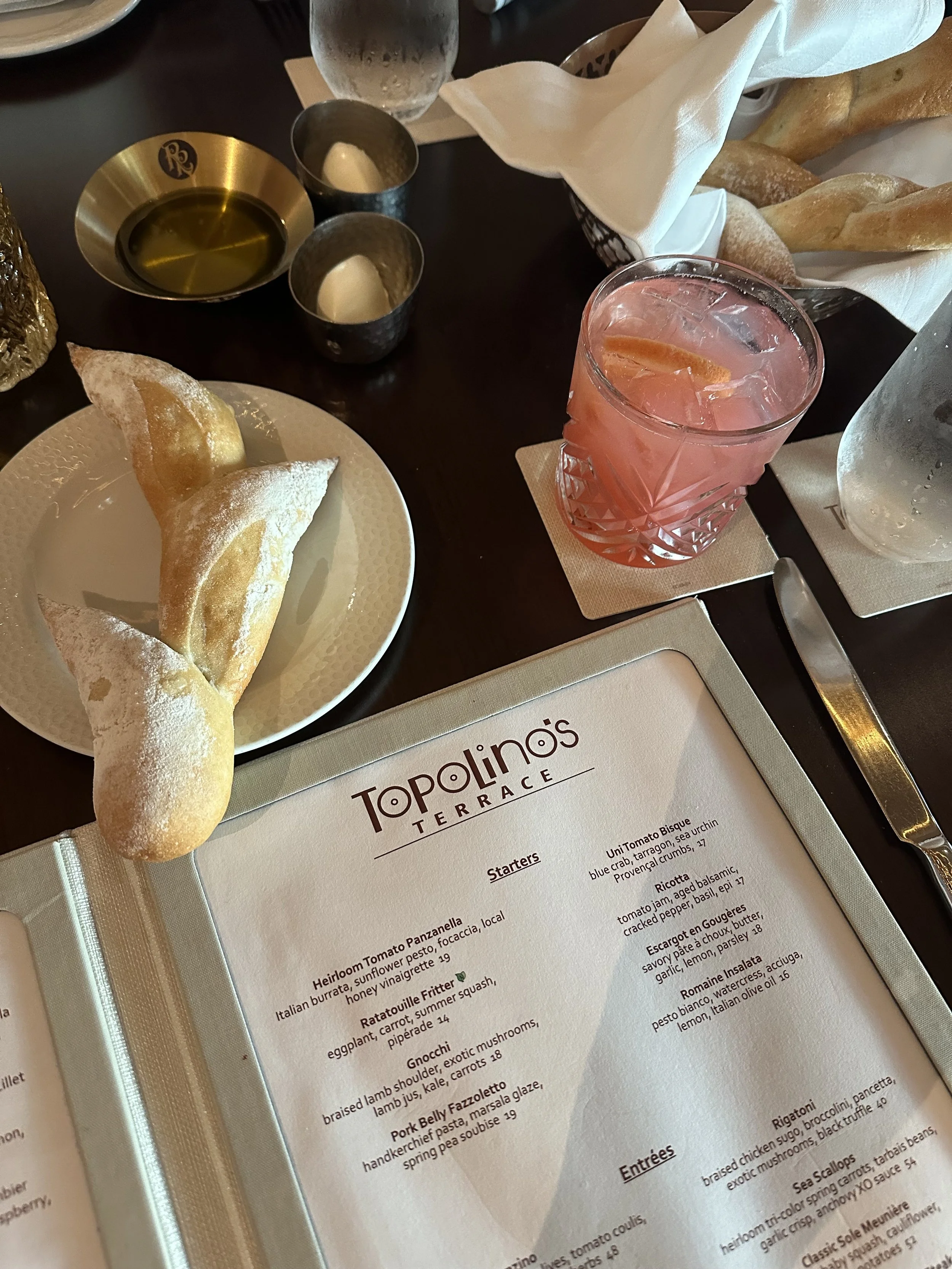 A restaurant table setting with a menu from Topolino's Terrace, a pink drink with ice, a basket of breadsticks, and a plate of three pastry turnovers dusted with powdered sugar.
