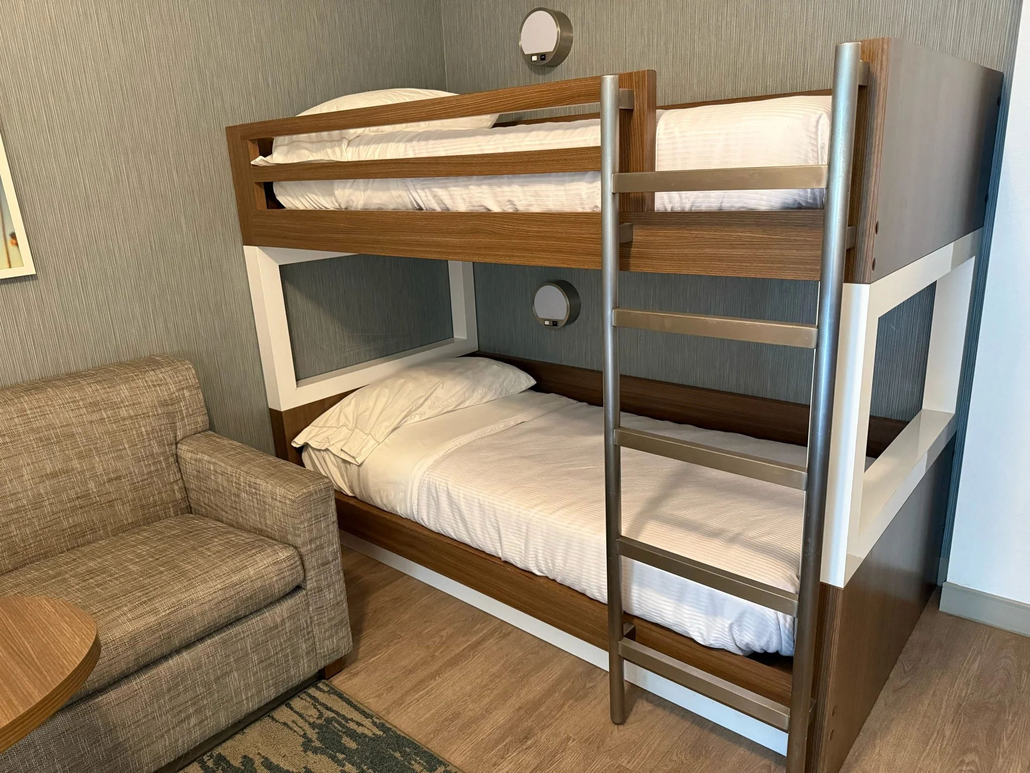 A bunk bed with a lower bed and an upper bed, both with white bedding and pillows, in a room with beige and brown decor. There is a beige plaid sofa nearby and a small round wooden table.