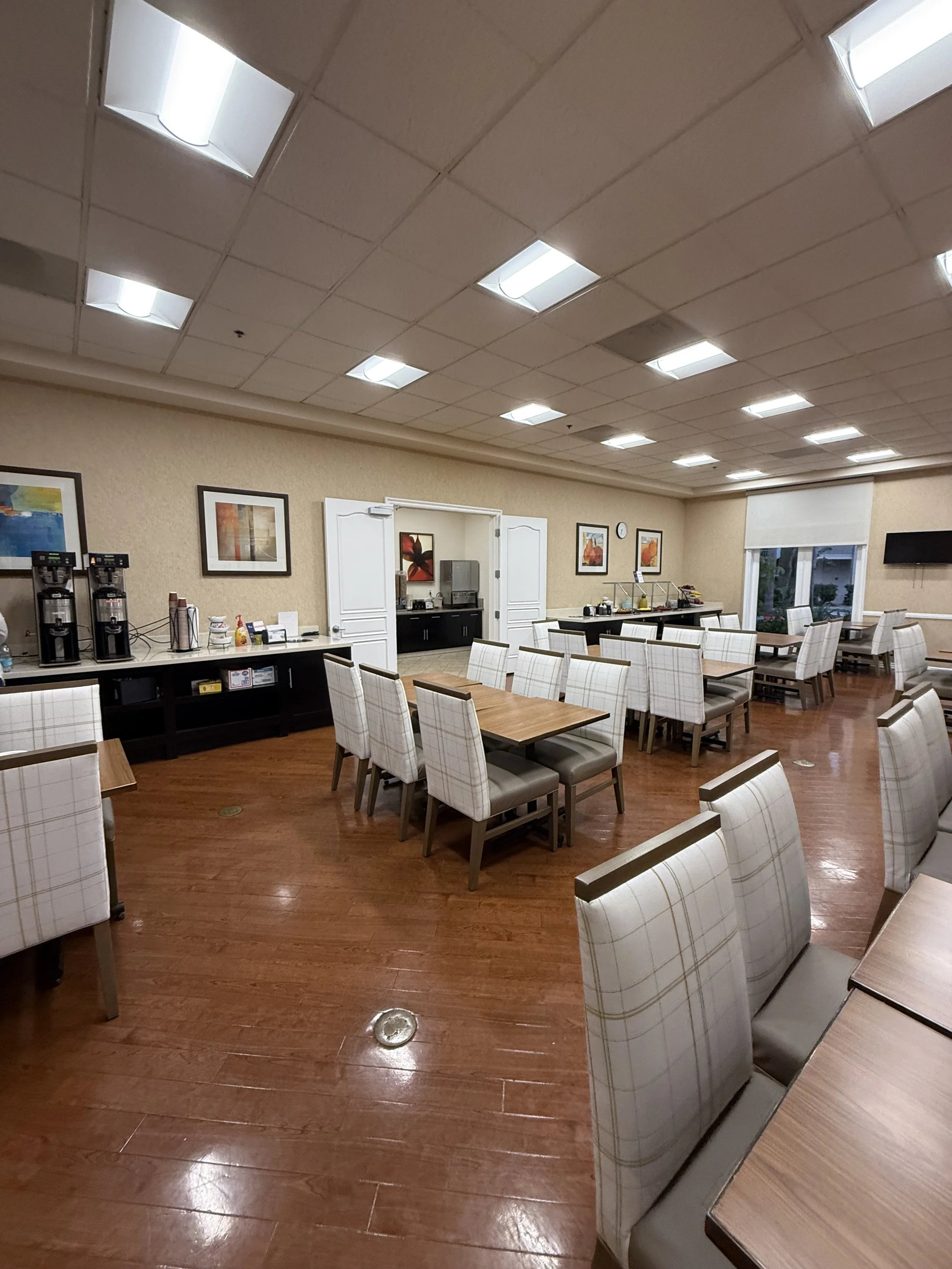 Empty dining area with wooden tables and plaid upholstered chairs, ceiling lights, artwork on beige walls, coffee station on left side, windows with blinds on the right, and buffet station against wall.