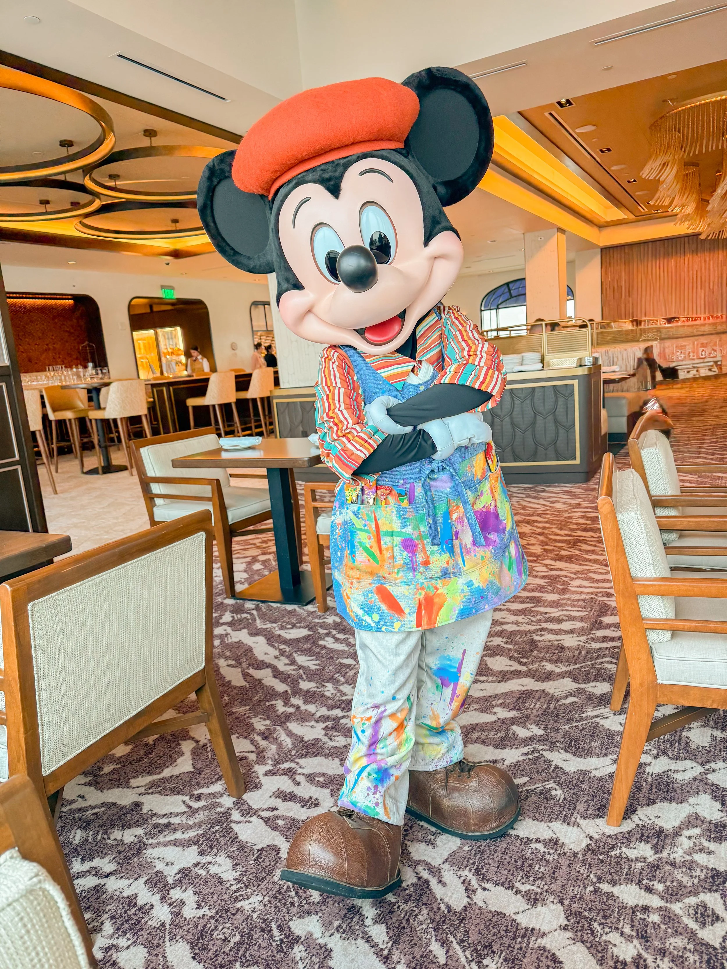 Costumed character of Mickey Mouse dressed in paint-splattered clothes and a beret, standing inside a restaurant.