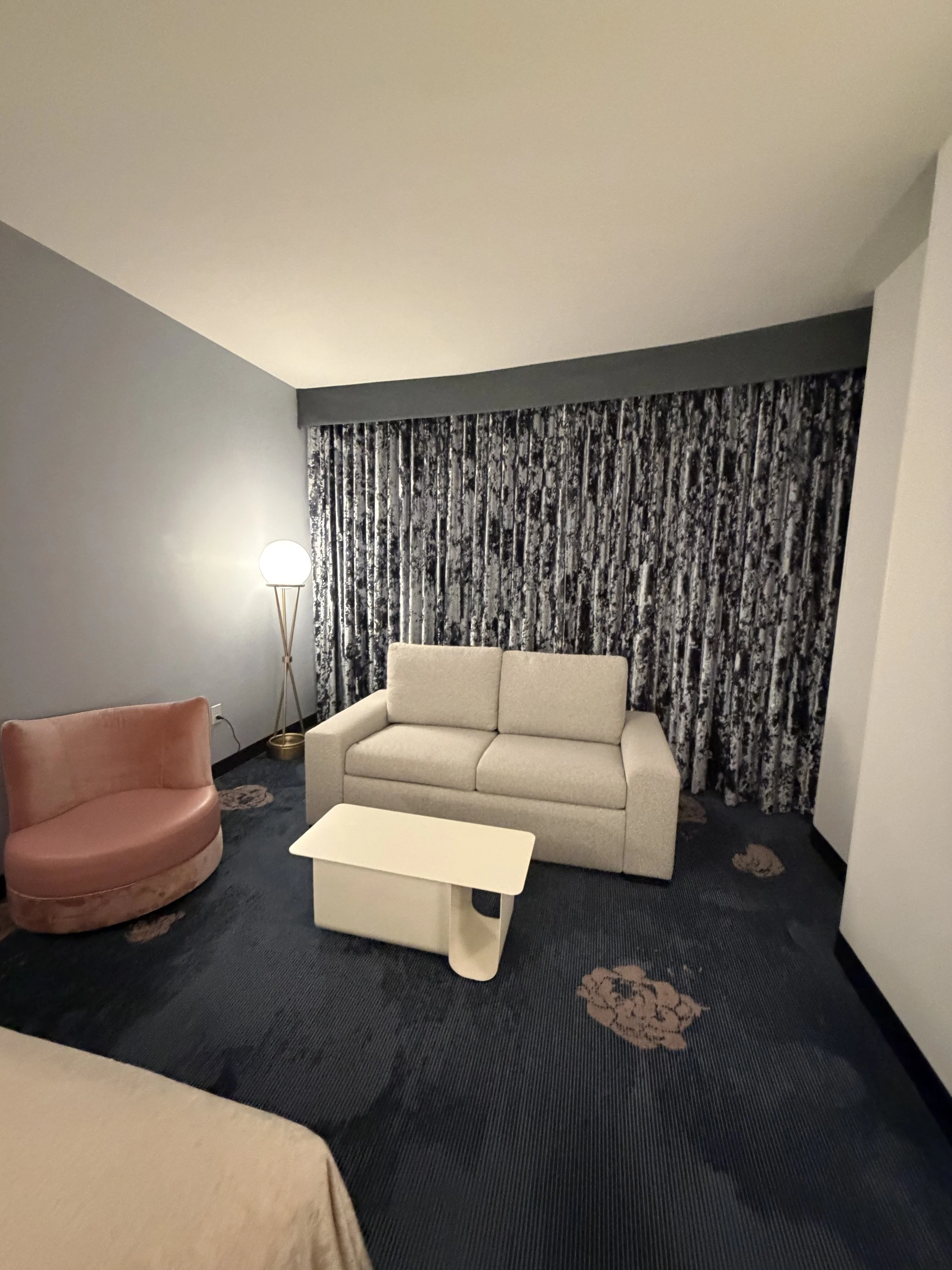 A hotel room with grey walls, a white sofa, a pink armchair, a white coffee table, a floor lamp, curtains with black and white pattern, and a carpet with floral design.