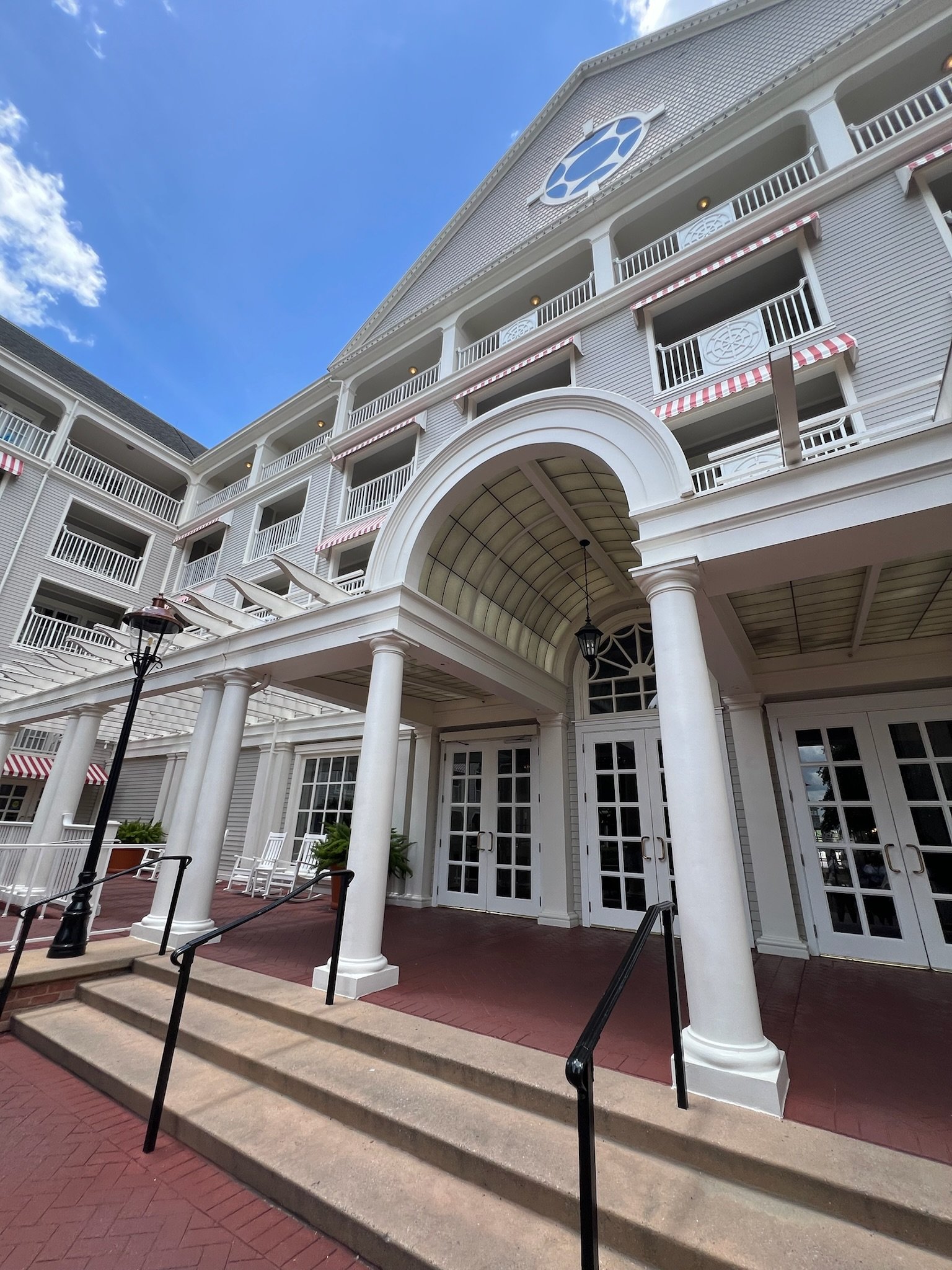 Exterior of a large, white, multi-story building with balconies that have red and white striped awnings outside each. The entrance has a large arched canopy with columns, glass doors, and potted plants, set on a brick walkway with steps and black met