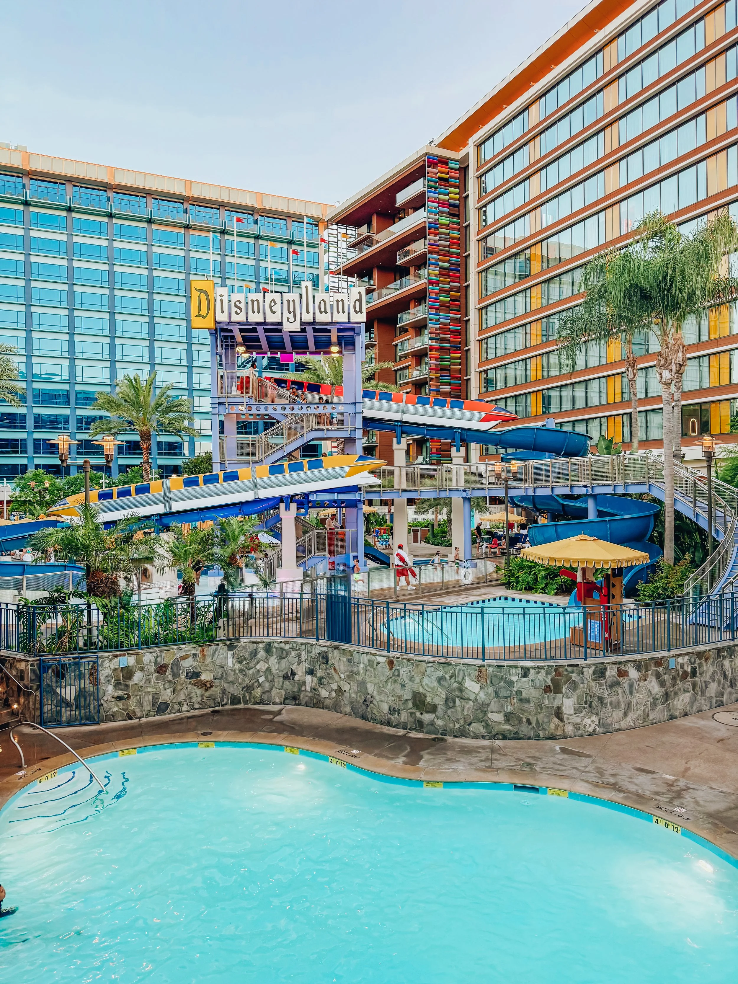 Water slide area at Disneyland hotel with blue and red slides, pool, and surrounding buildings with glass windows and palm trees.