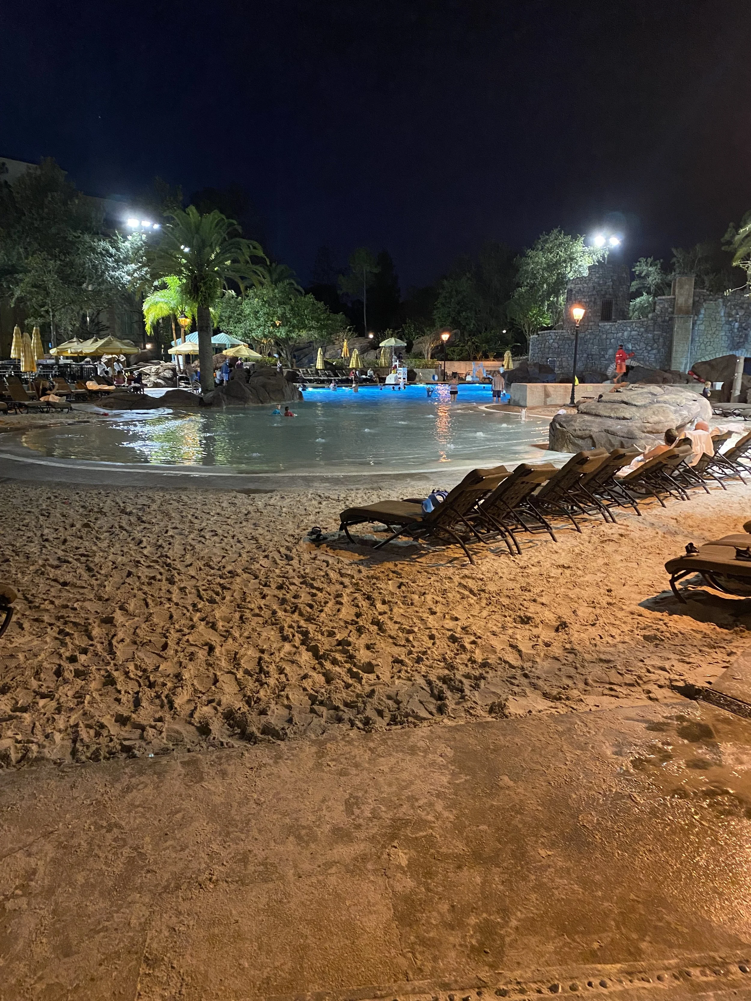 Night view of a swimming pool area with lounge chairs, umbrellas, and people swimming, surrounded by trees and rocks, with lights illuminating the scene.