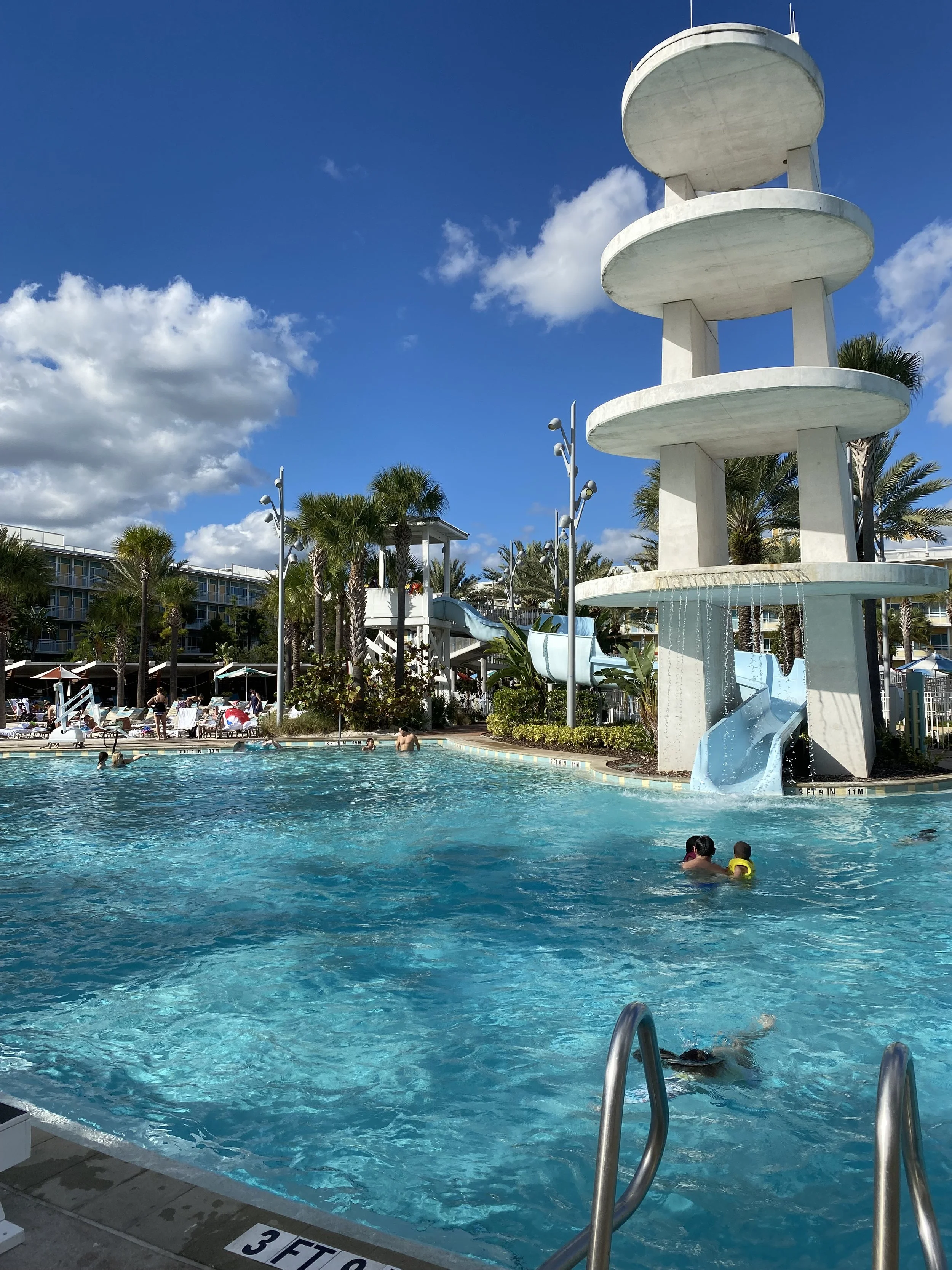 Aquatic pool with a water slide and tower at a resort, surrounded by lounge chairs and palm trees under a blue sky.
