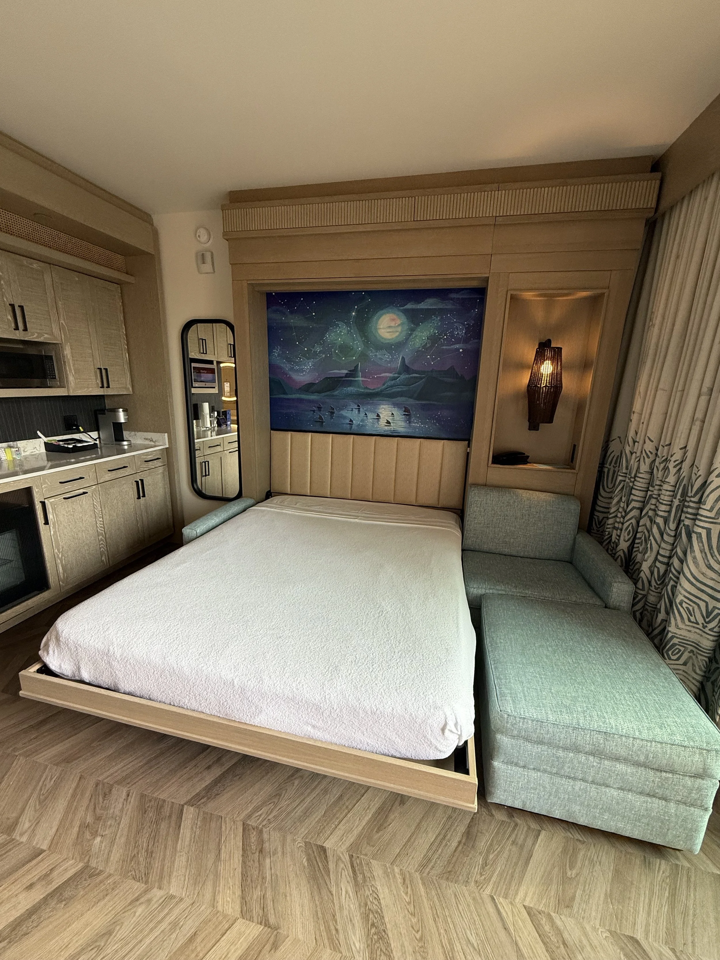 Hotel room with a bed, artwork of an aurora borealis and mountains, a sofa, a lamp, and a kitchenette area.