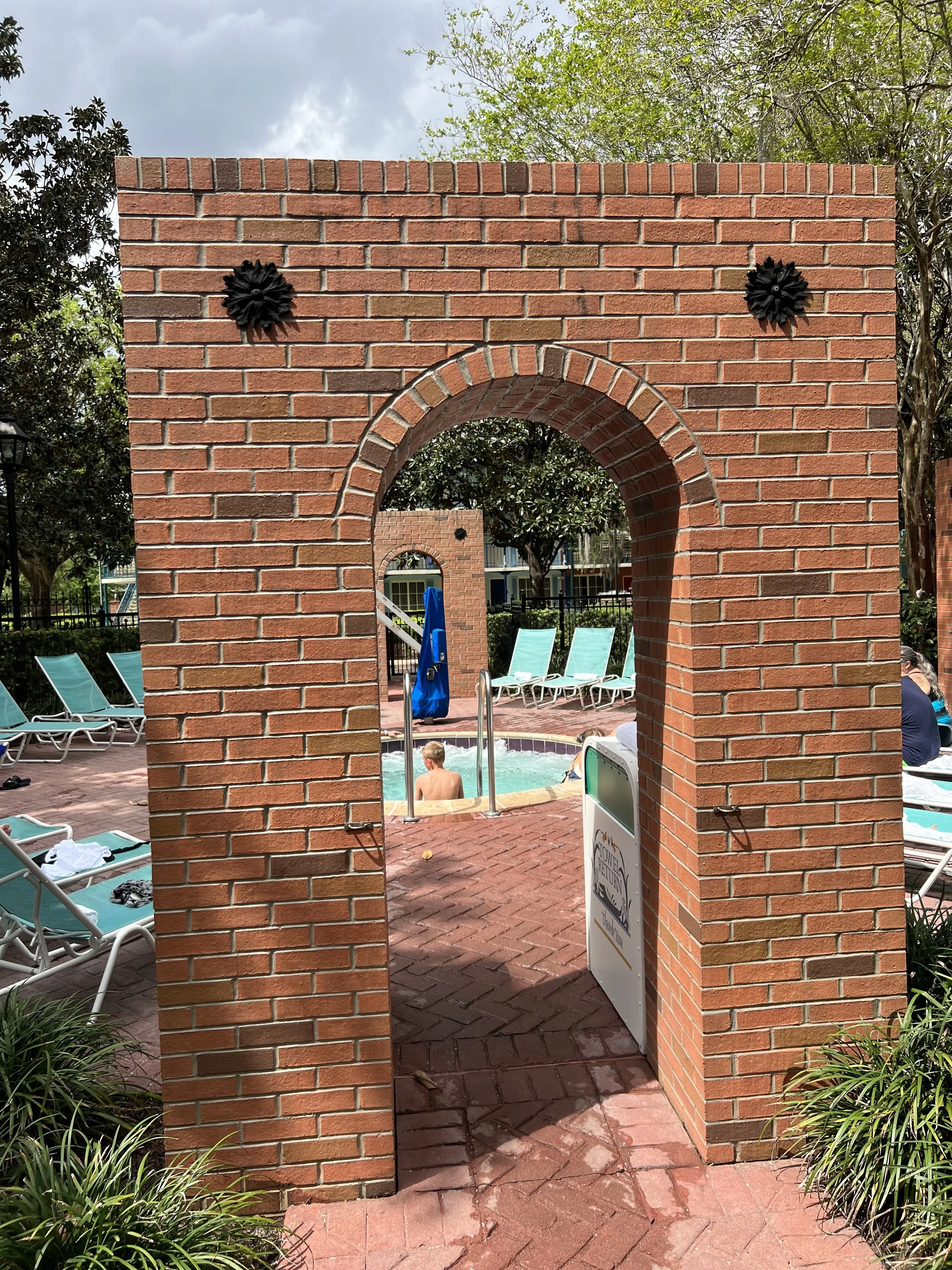 View of a swimming pool area through a brick archway, with lounge chairs around the pool and a boy in the water, surrounded by trees and umbrellas.