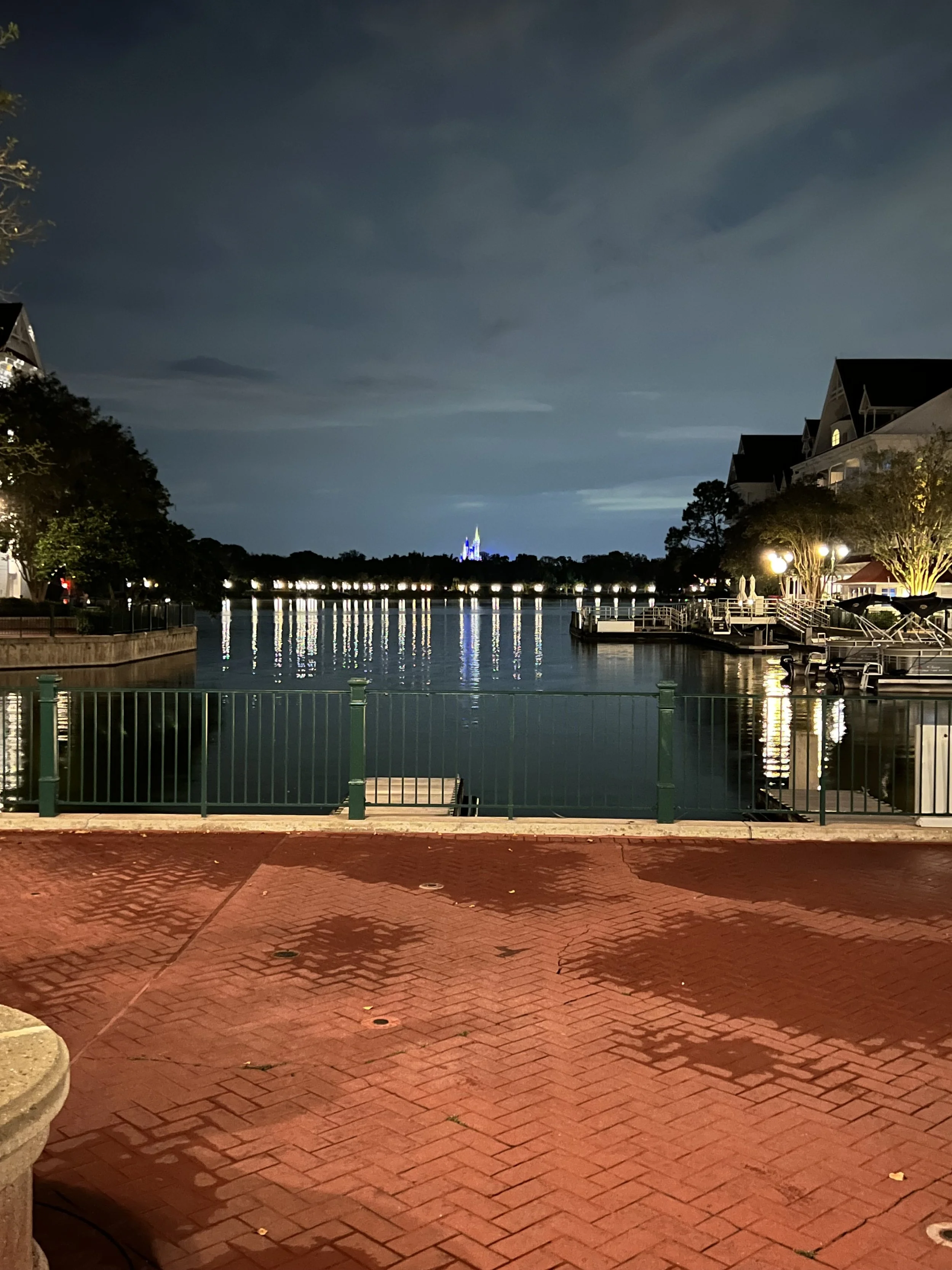 Nighttime view of a lake or harbor with calm water reflecting lights from surrounding buildings. In the background, a lit-up castle or tower can be seen, resembling Disney Castle. The foreground features a brick-paved area and a green railing along t