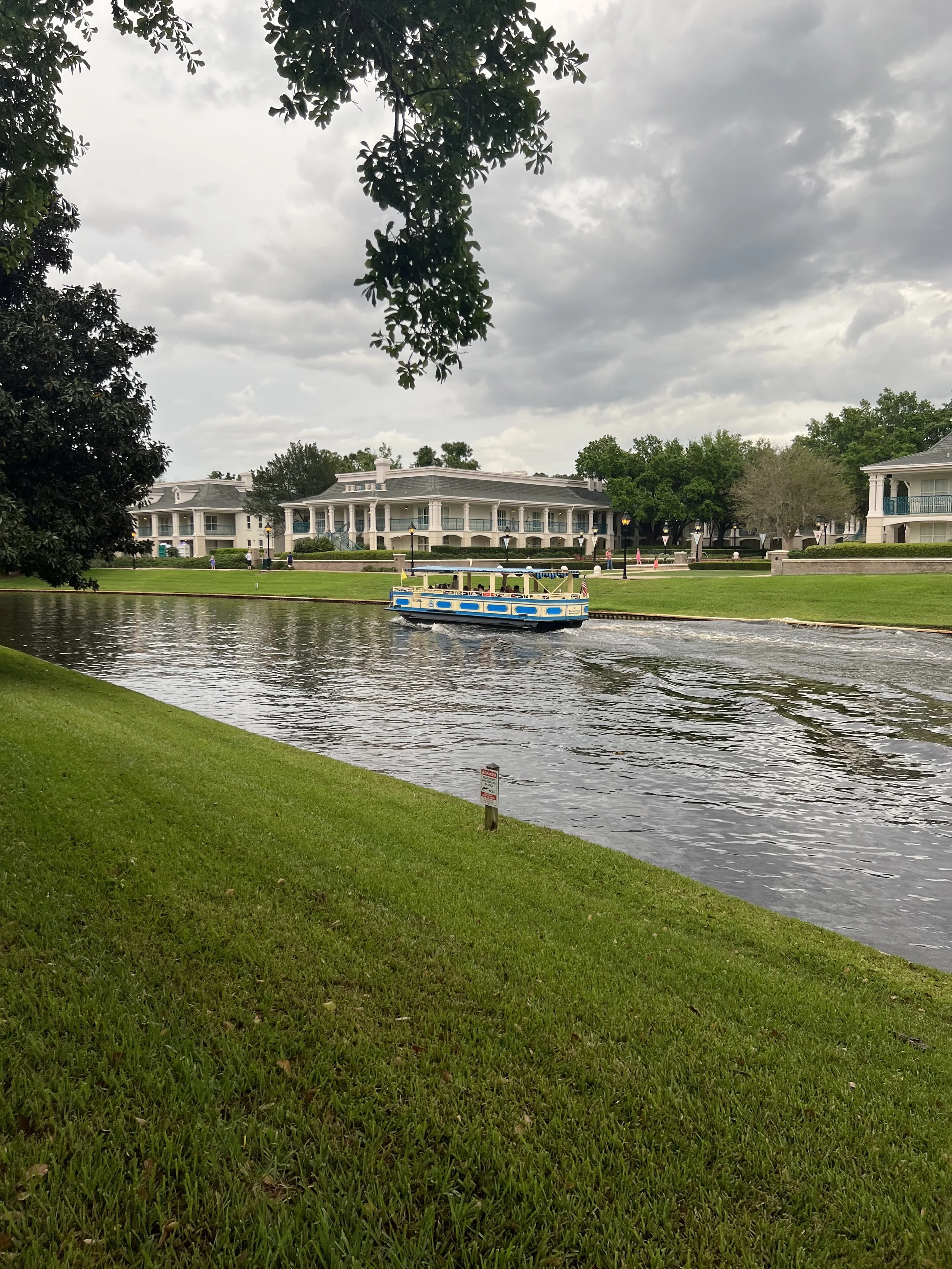 A boat on a small lake surrounded by green grass and trees, with a row of houses in the background under a cloudy sky.