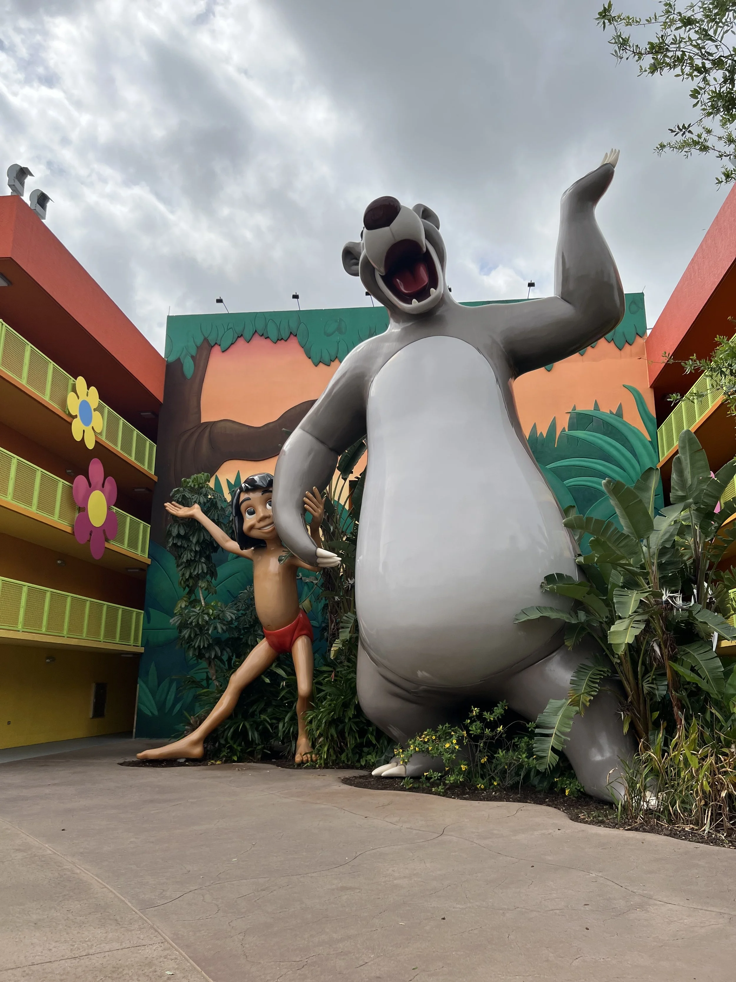 Large cartoon statues of Baloo the bear and Mowgli the boy from The Jungle Book, standing outdoors near greenery and a colorful building with yellow balconies and flower decorations.