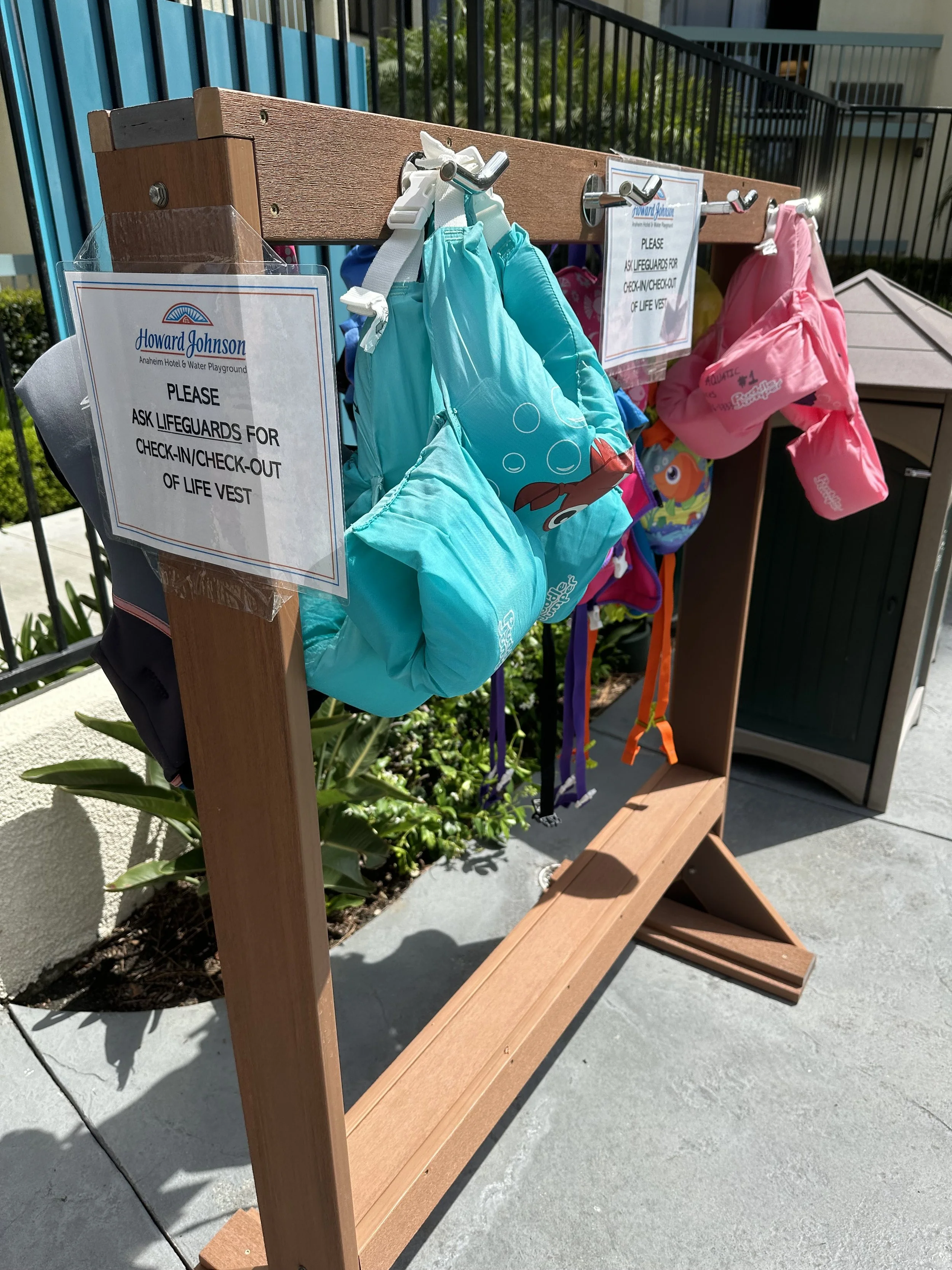 Outdoor lifeguard safety station with backpacks and pink life jackets hanging on a wooden frame, with a sign requesting guardians to check life vests.