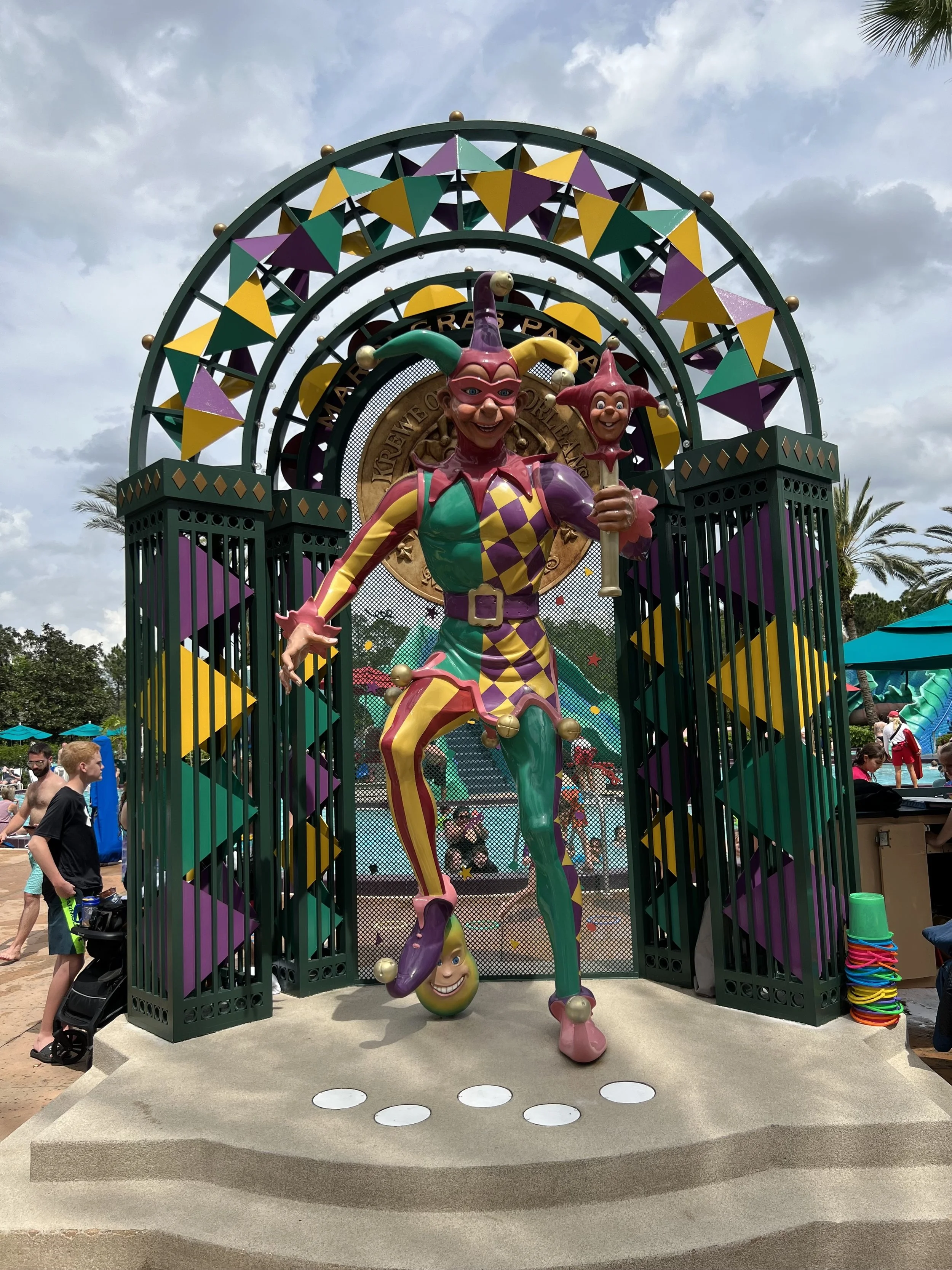 Colorful jester figure at a carnival or amusement park with an archway in the background and people nearby.