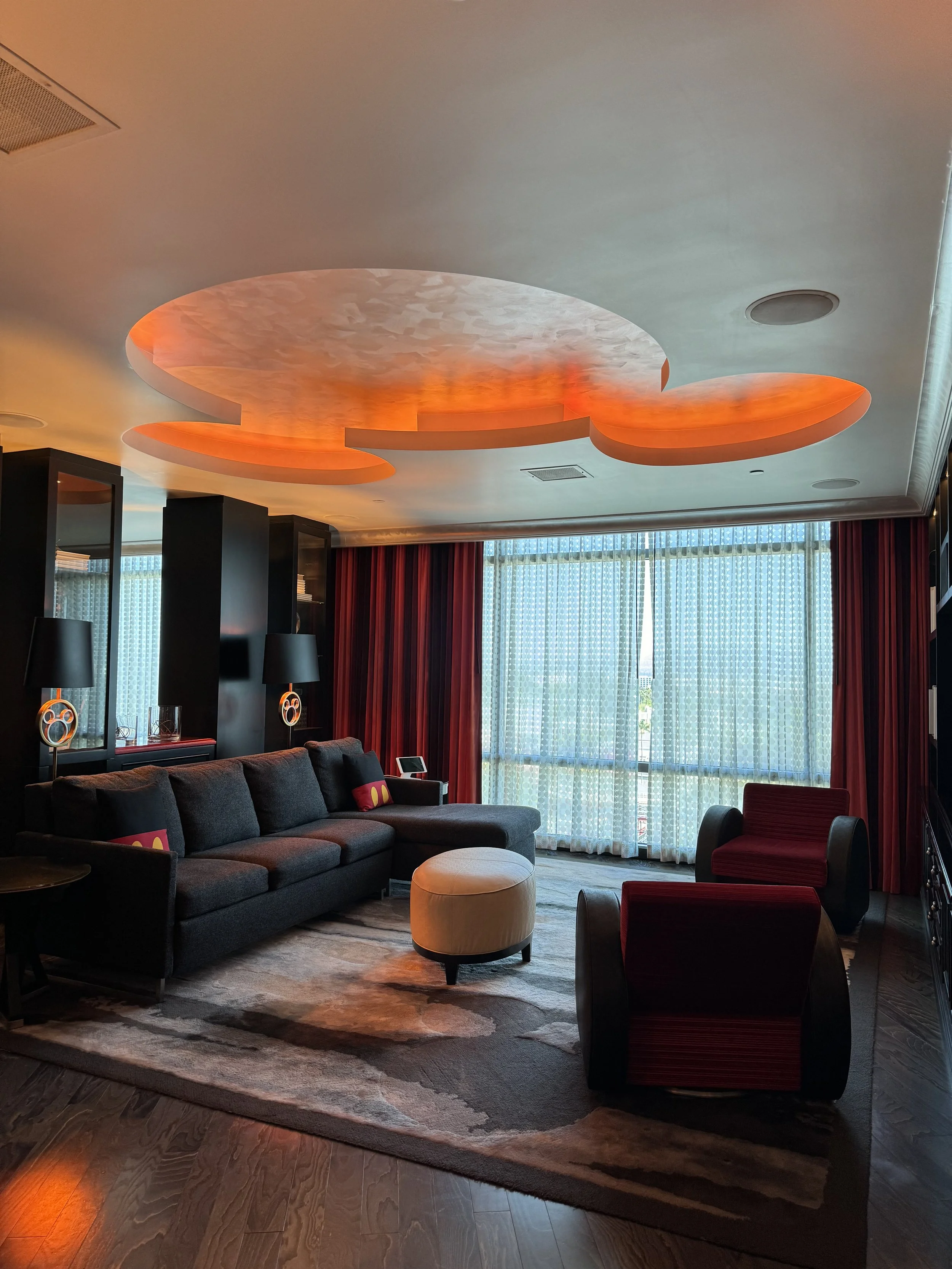 Modern hotel living room with dark gray sofa, two red and black armchairs, white ottoman, patterned area rug, black side table, and large windows with sheer white curtains and red drapes. Unique orange-lit, cloud-shaped ceiling fixture.
