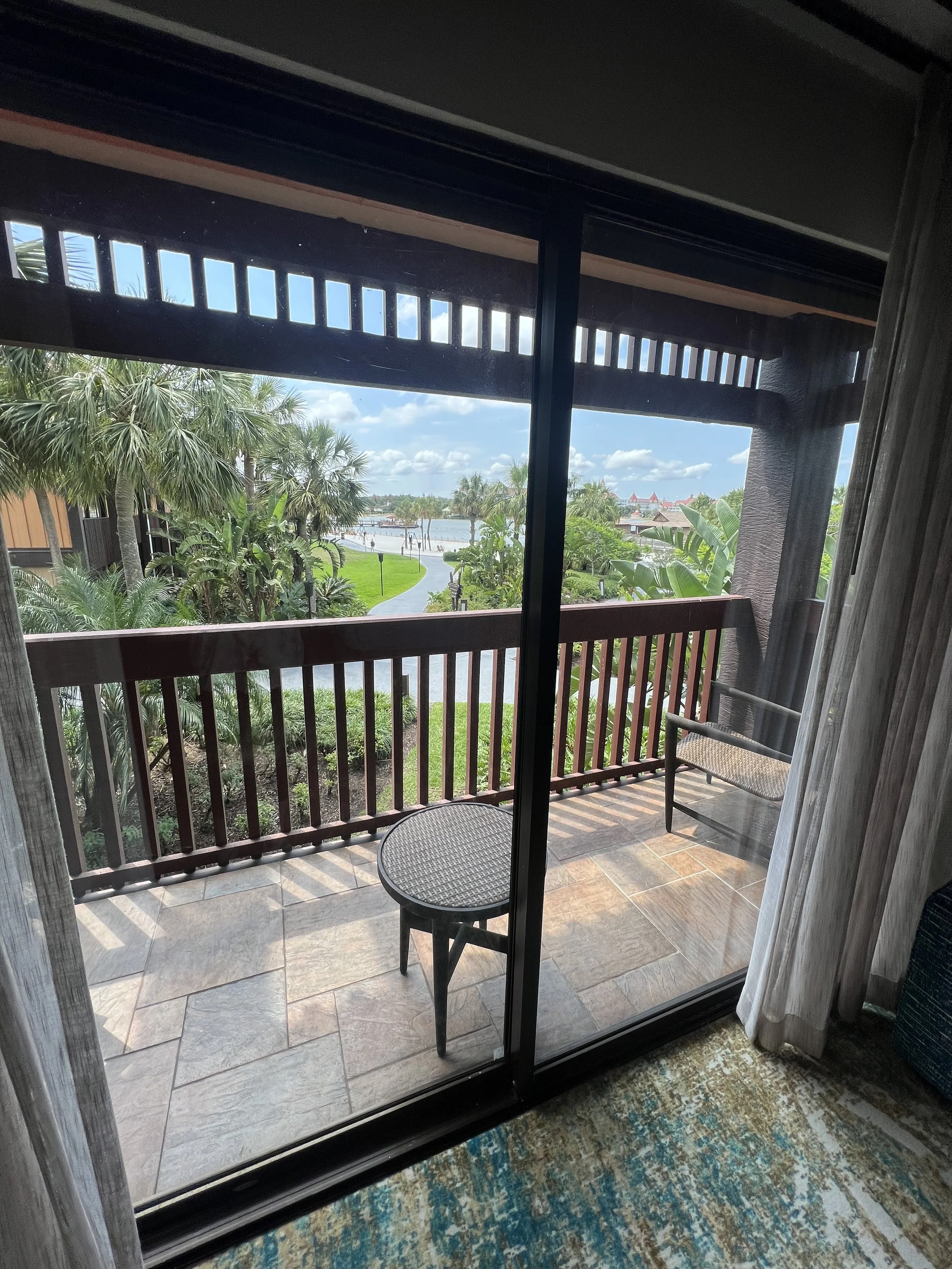 View of a balcony with a wooden railing, a small round table, and a chair, overlooking a lush green landscape with palm trees, a pathway, water, and buildings in the distance under a partly cloudy sky.