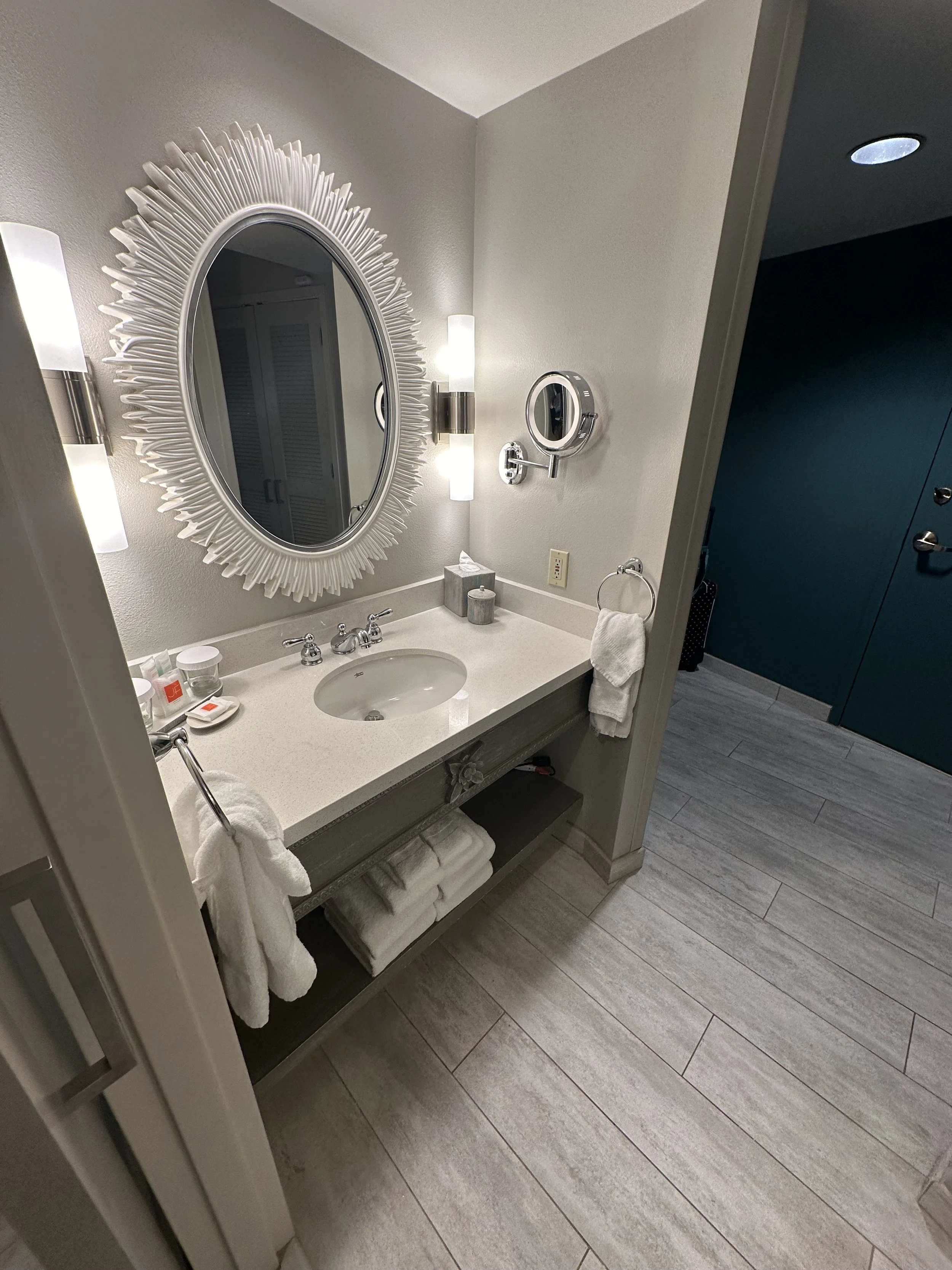 A bathroom vanity with a large decorative mirror, two wall sconces, and a mounted vintage-style magnifying mirror. The vanity has a soap dish, tissue box, and toiletries on the countertop, with towels stored on the open shelf below. The floor is wood