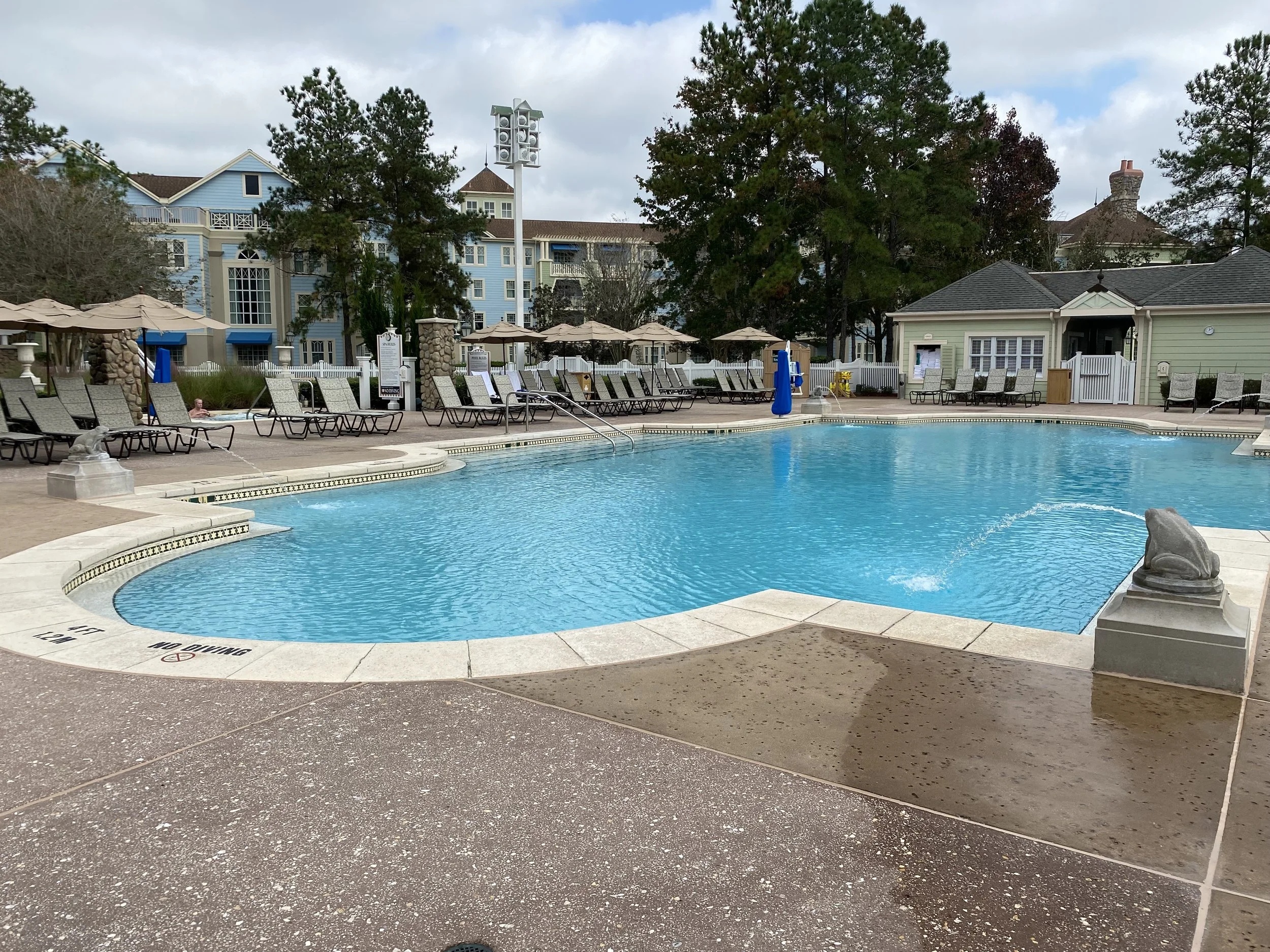 Empty outdoor swimming pool with lounge chairs, umbrellas, and a small building in a residential area, surrounded by trees and multi-story buildings in the background.