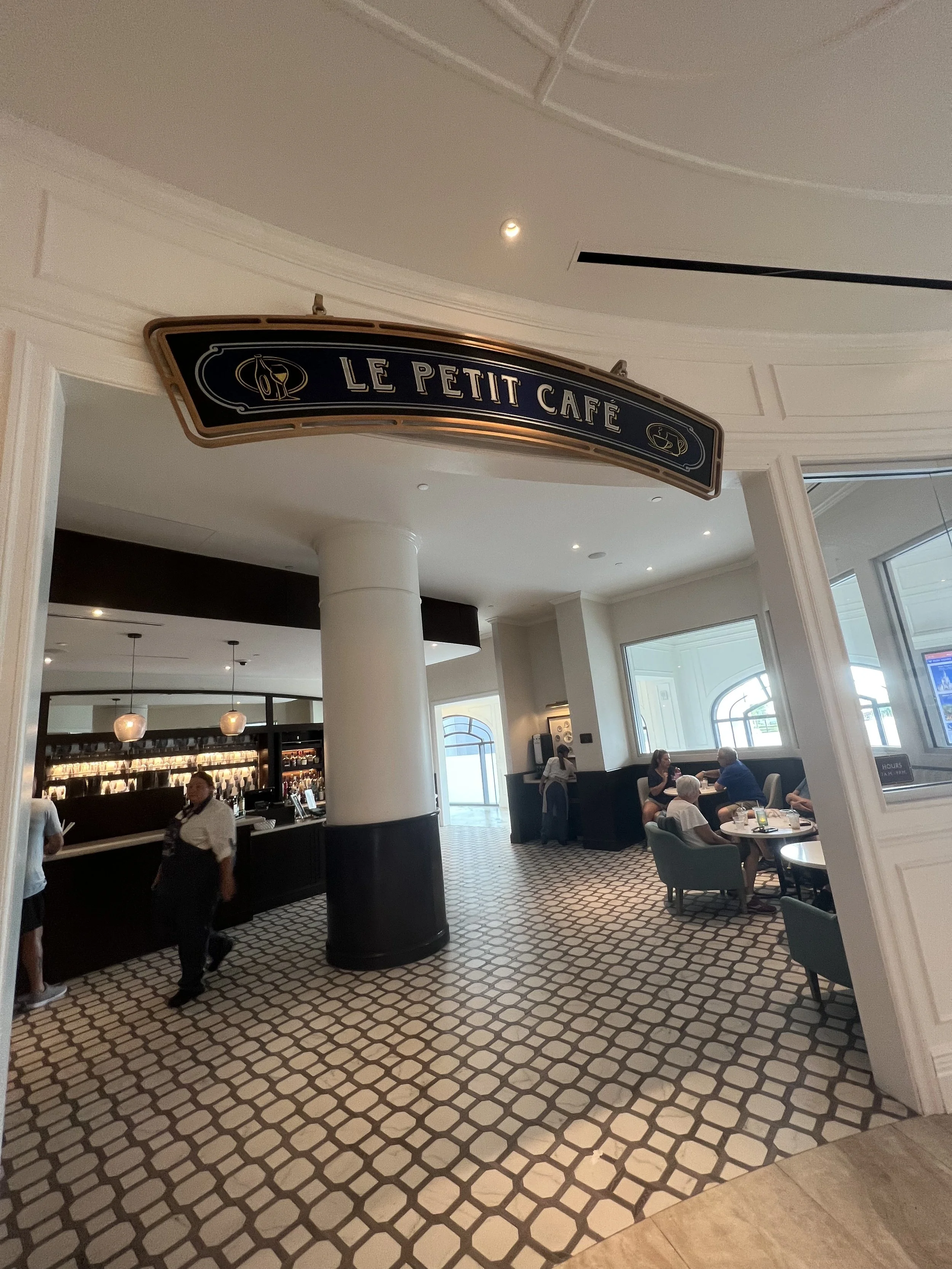 Interior of Le Petit Café with a curved entrance, a patterned tile floor, a small bar area, and tables with patrons dining. Sign with the café’s name hanging above the entrance.