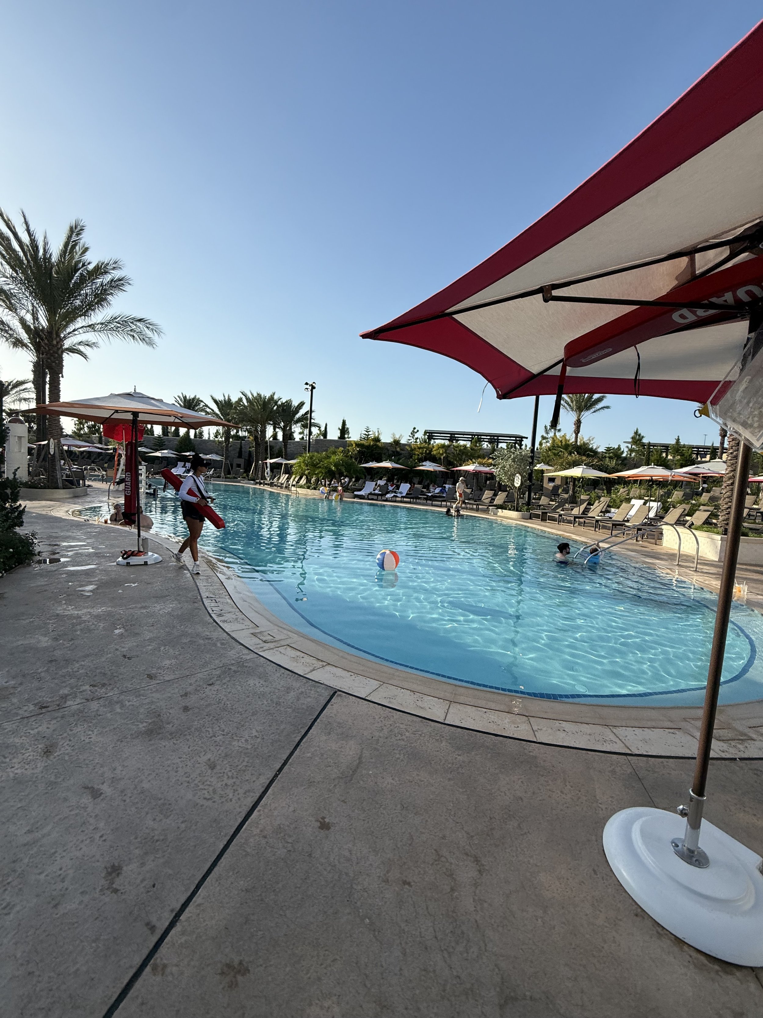 Poolside scene with a swimming pool, lounge chairs, umbrellas, palm trees, and people swimming and relaxing.