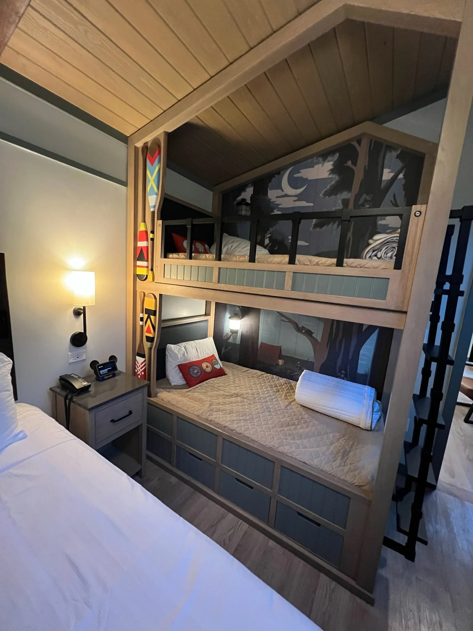 A hotel room with a bunk bed that has a forest and night sky mural on the headboard panel, a single bed with white sheets, a nightstand with a phone and alarm clock, and two decorative paddles hanging on the wall.