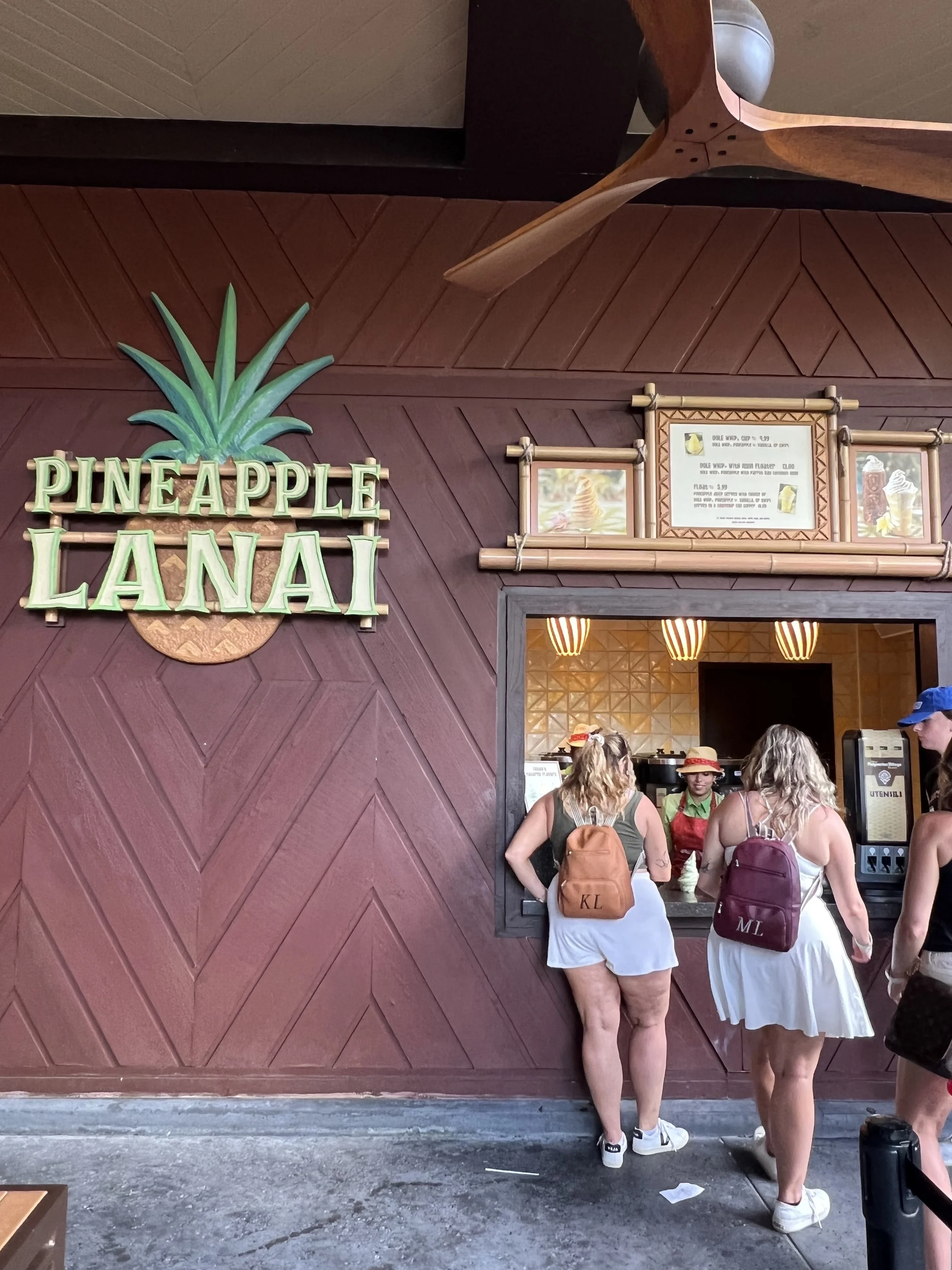 People ordering food at the Pineapple Lanai restaurant counter with a sign featuring a pineapple and tropical decor.