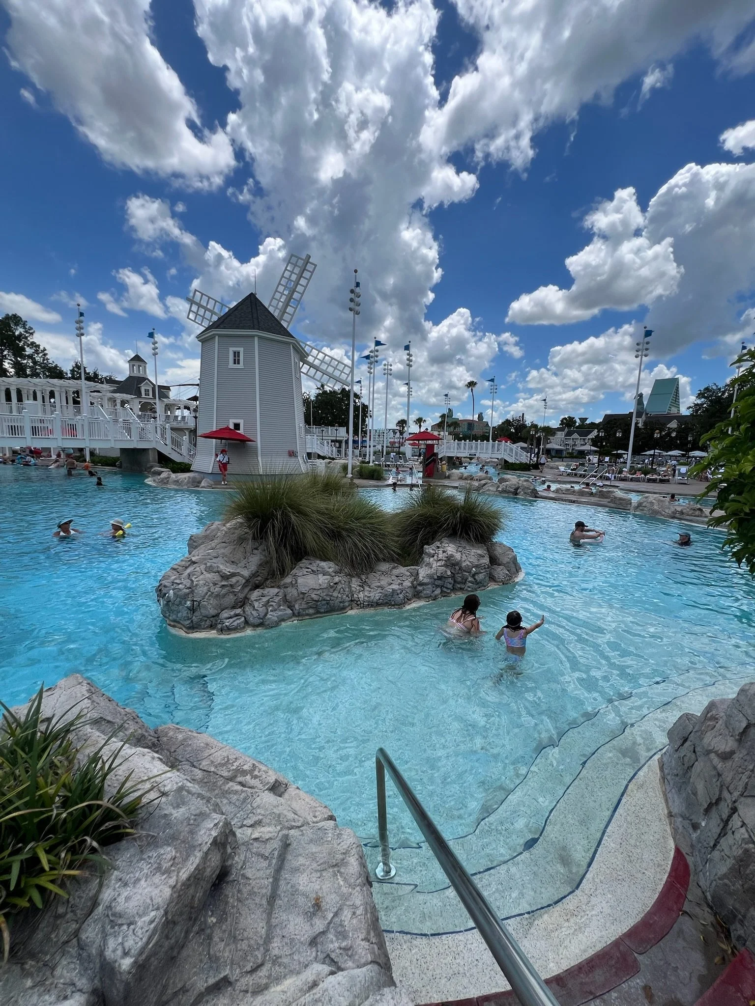 People swimming and relaxing in a large outdoor pool with a mini windmill, rocks, and plants, under a partly cloudy sky.