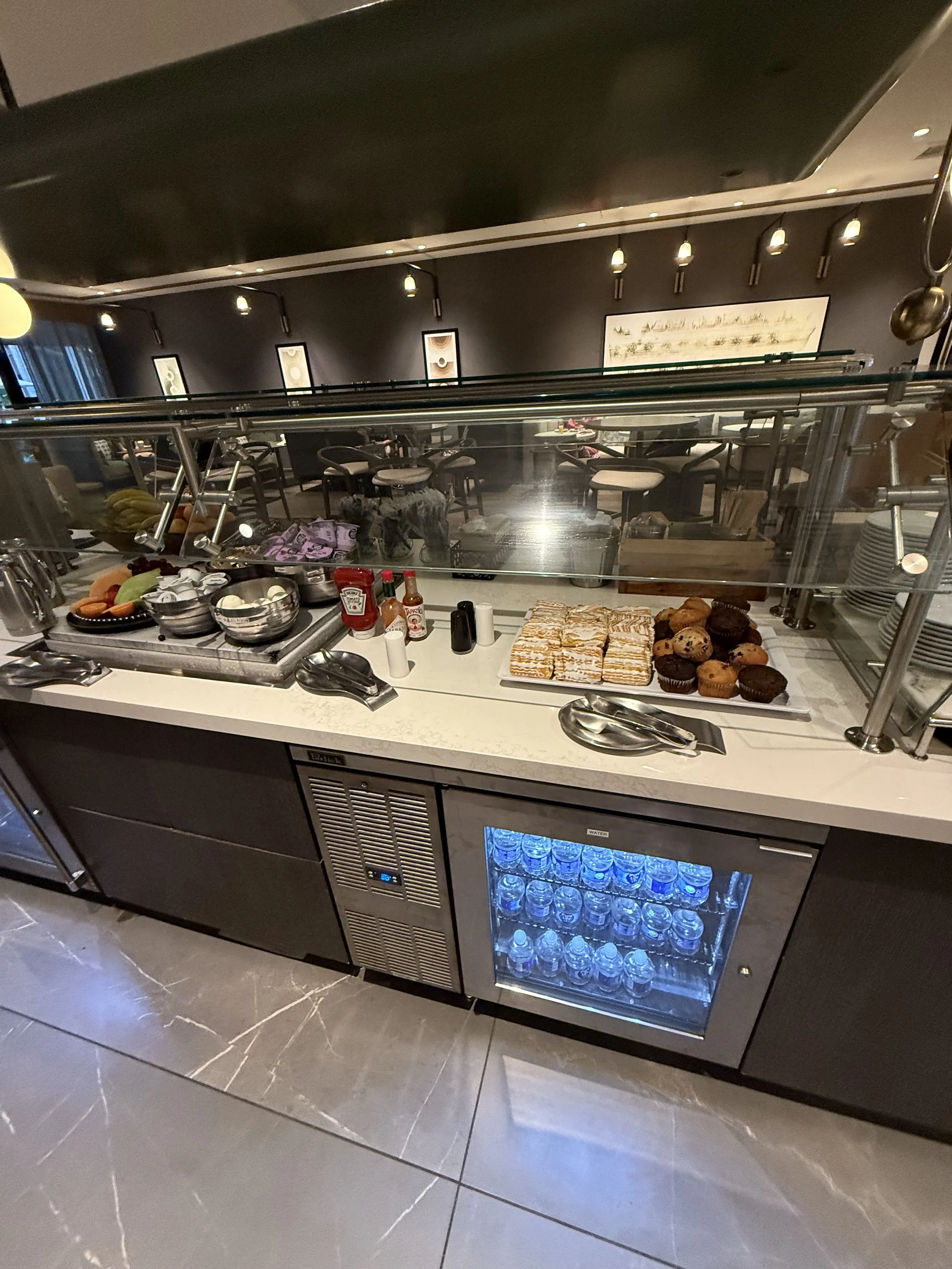 Buffet area with condiments, baked goods, and a mini fridge with water bottles in a restaurant or lounge setting.