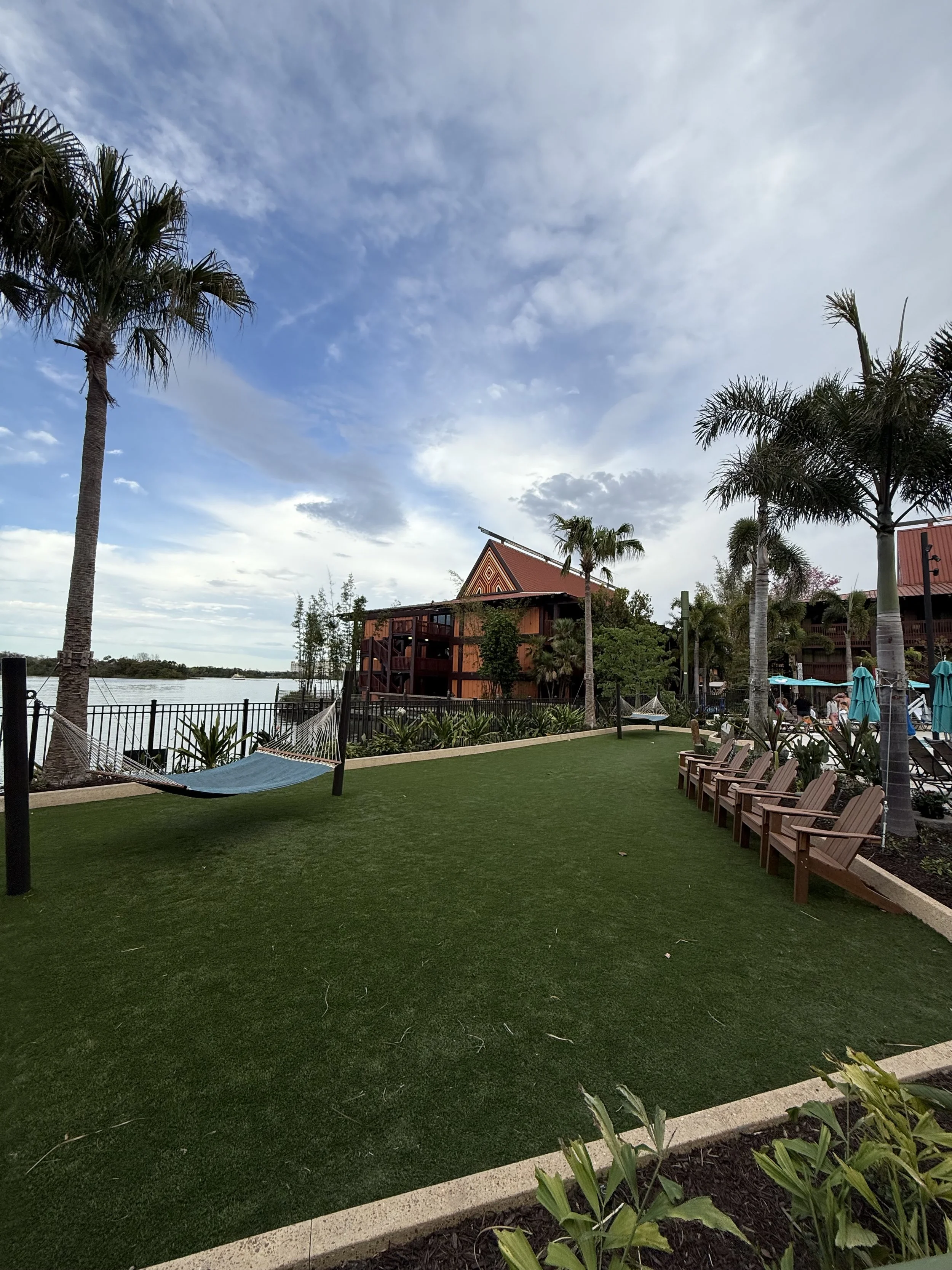 Lawn area with palm trees, lounge chairs, hammocks near a water body, and a building with a red roof under a partly cloudy sky.