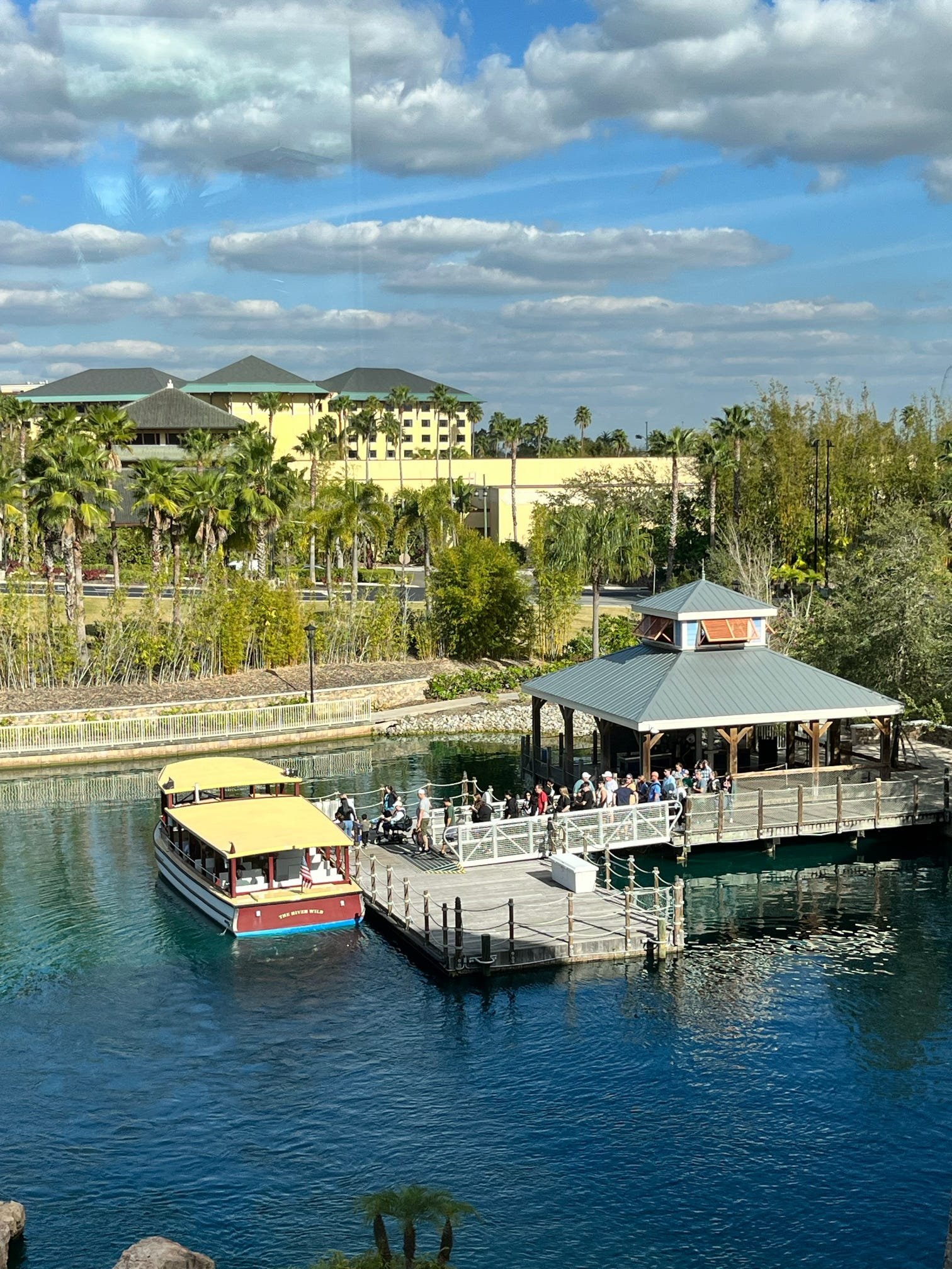 People waiting on a dock next to a boat at a park with a pond, surrounded by trees and buildings, under a partly cloudy sky.