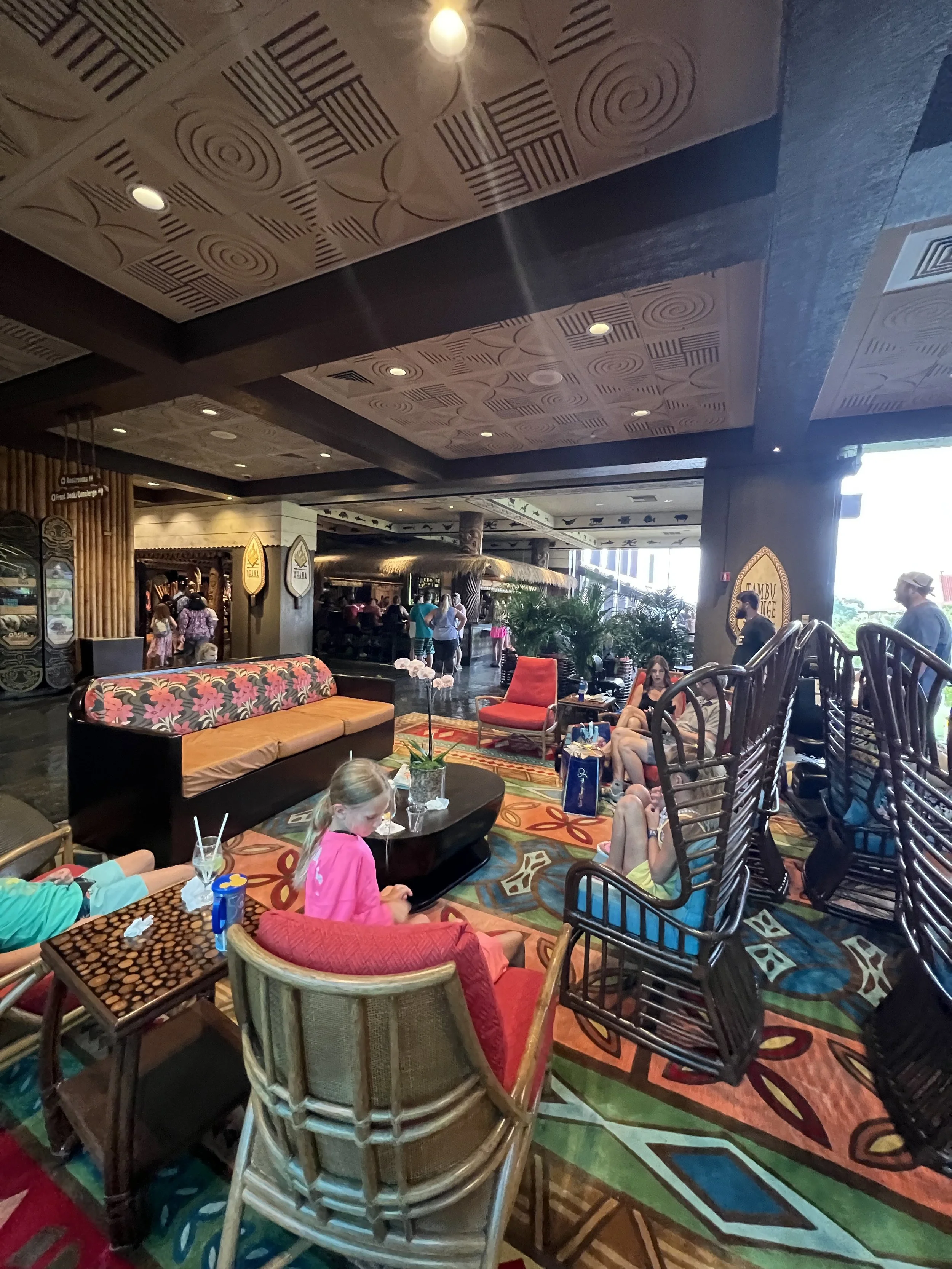 The image shows an indoor lounge area with colorful patterned carpet, various seating options including rattan chairs, a floral sofa, and a red armchair. Several children and adults are relaxing, socializing, or standing around. The ceiling has a dec