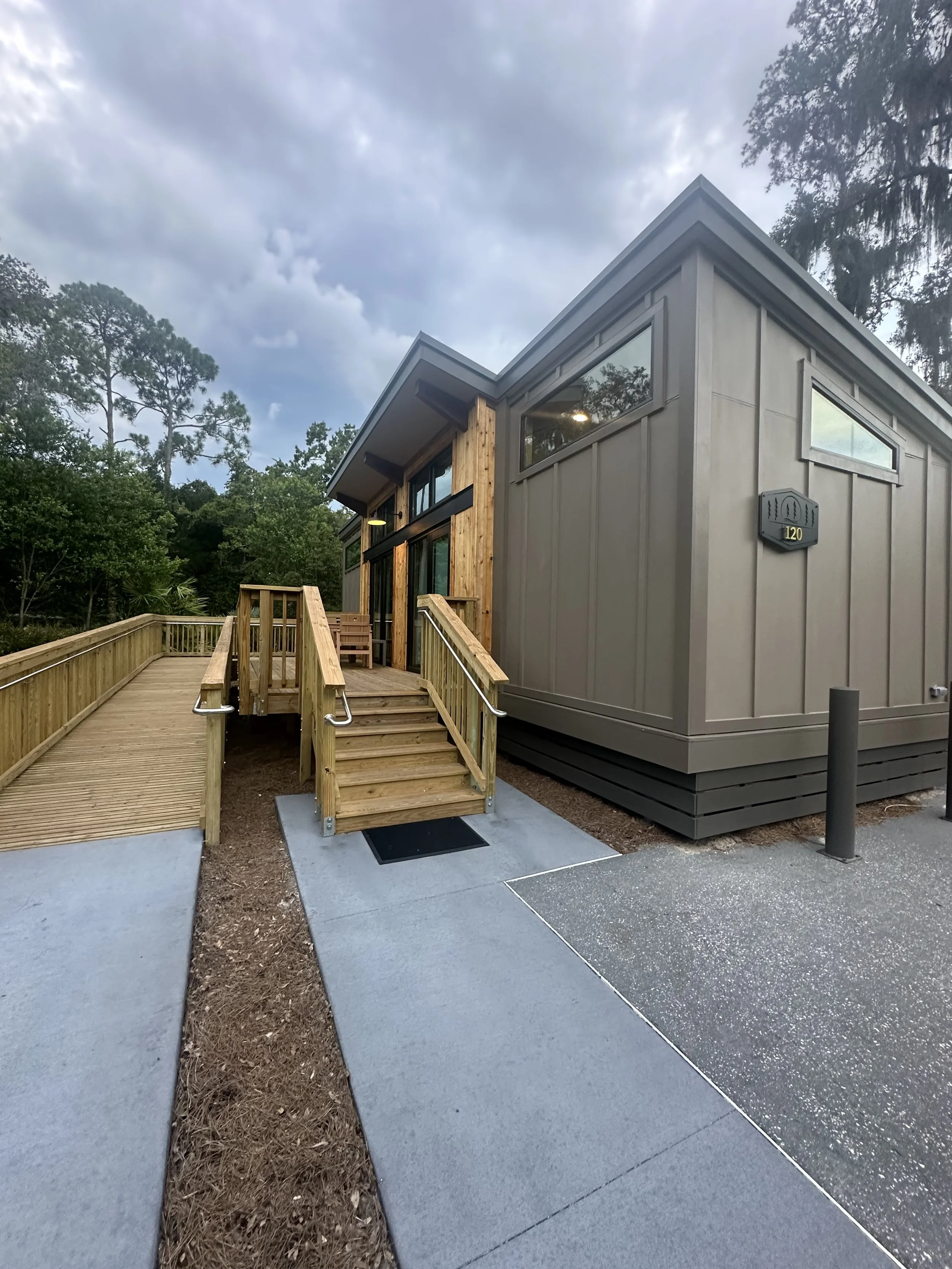 Front view of a modern house with a wooden ramp and stairs, gray exterior siding, and large windows, set against a backdrop of wooded trees and an overcast sky.