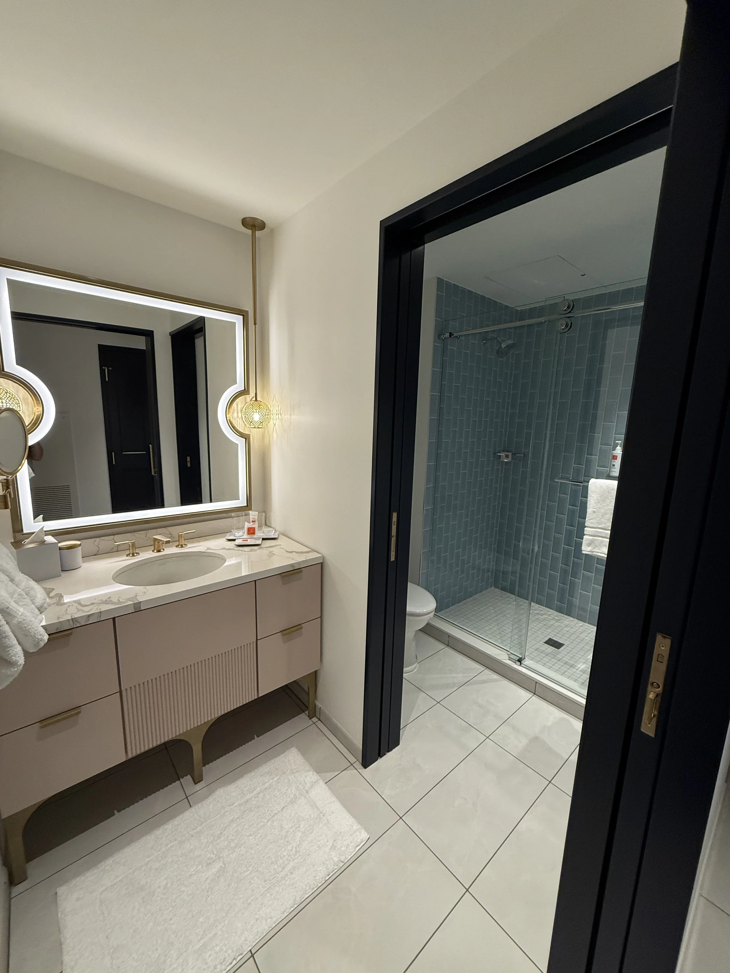 Modern bathroom with a vanity mirror, sink, and marble countertop. A hanging light fixture and a walk-in shower with blue tiles are visible.