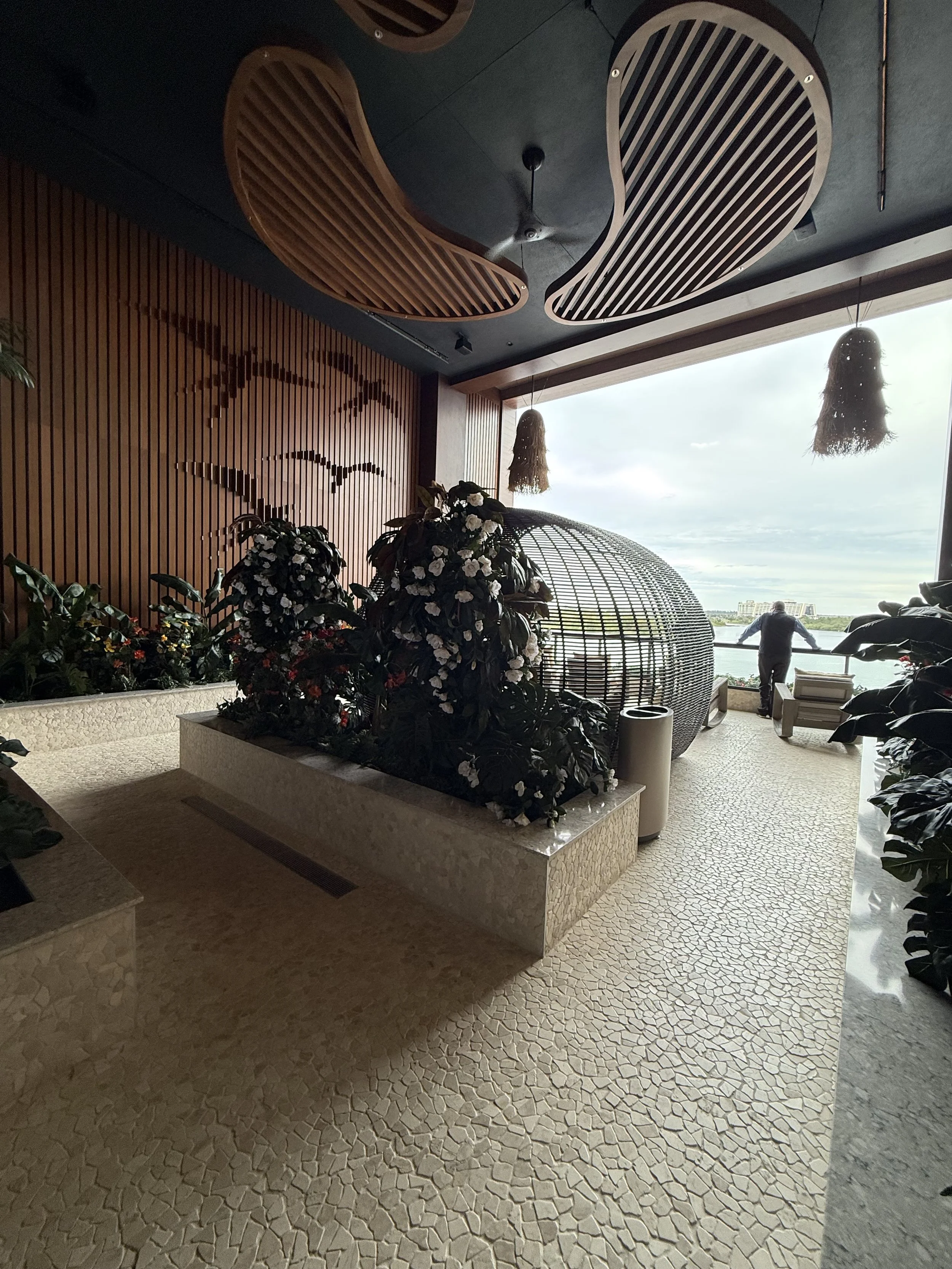 Indoor area with plants, decorative wooden ceiling panels, large window with view of water, and a person standing near the window.