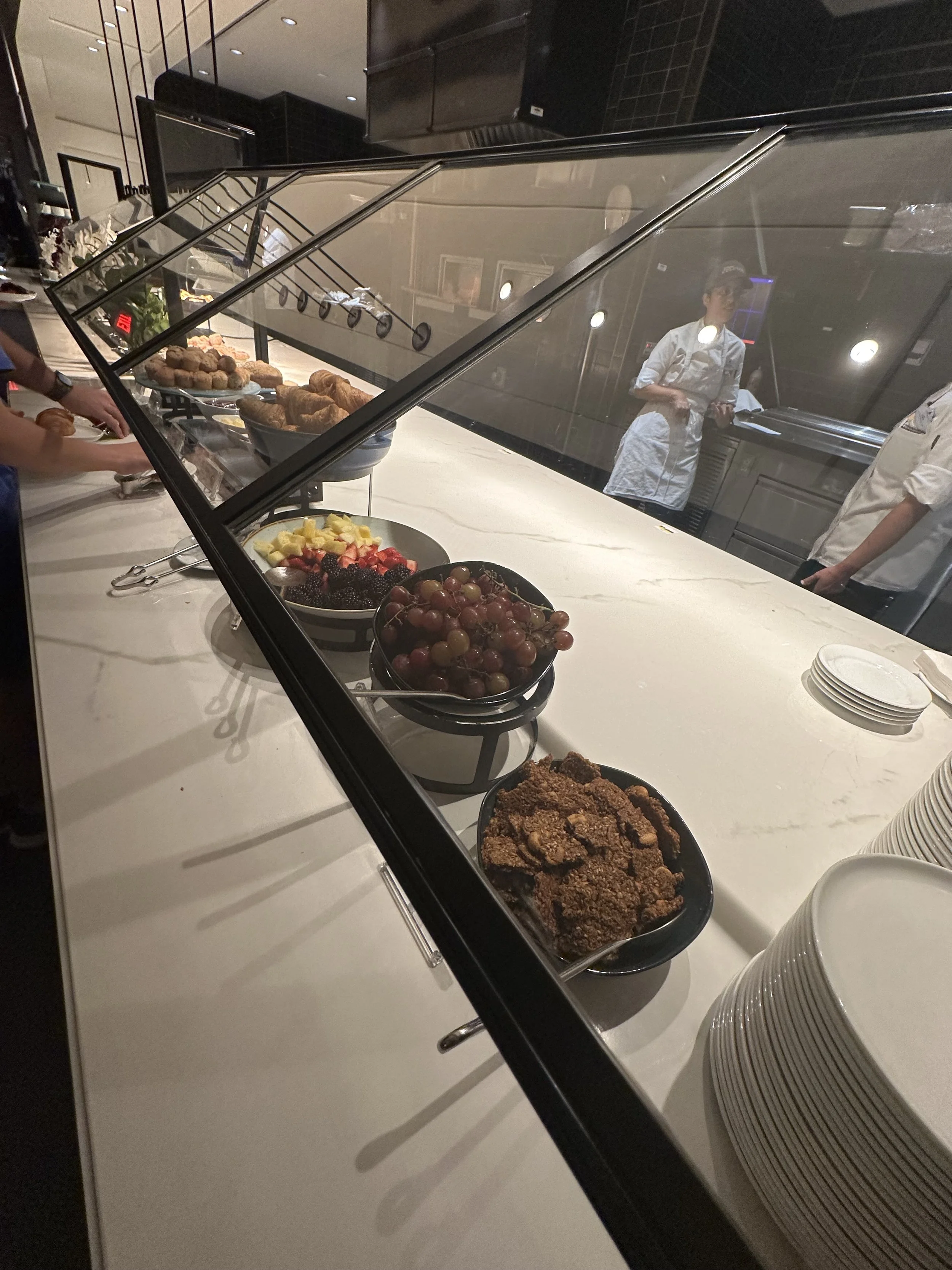 Buffet table with various breakfast foods such as fruit, pastries, and fried items, separated by a glass sneeze guard, with plates stacked nearby and staff members visible behind the counter.