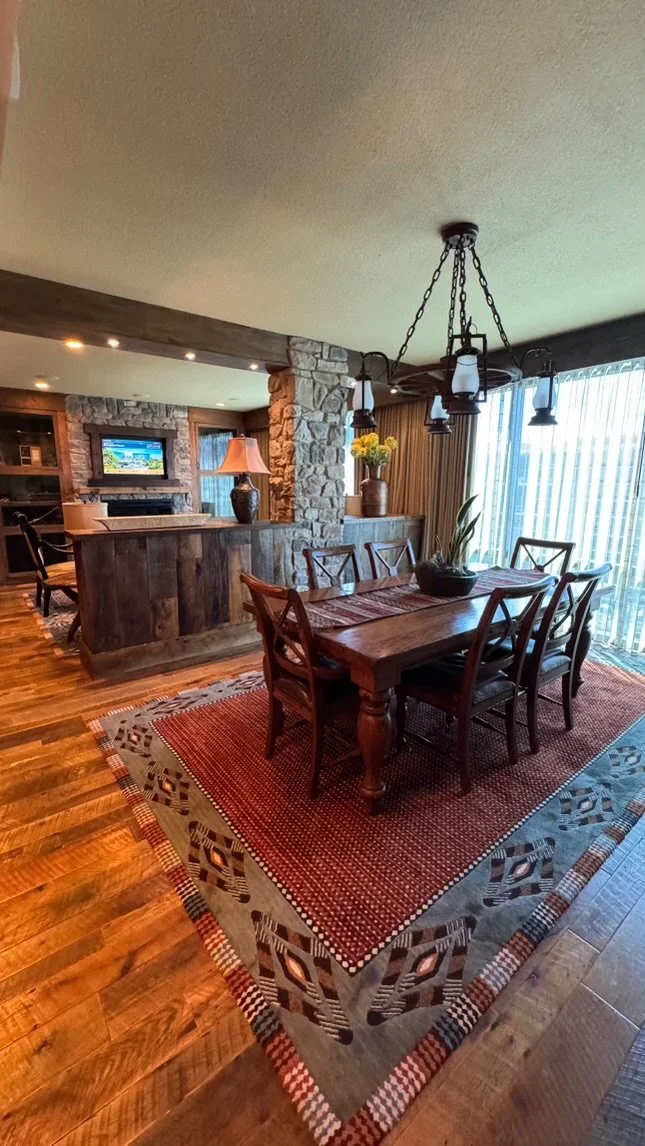 A dining room with a wooden table and six matching chairs, placed on a patterned area rug. The room features large windows with vertical blinds and a chandelier hanging above the table. There are decorative vases with flowers on a sideboard and a sto