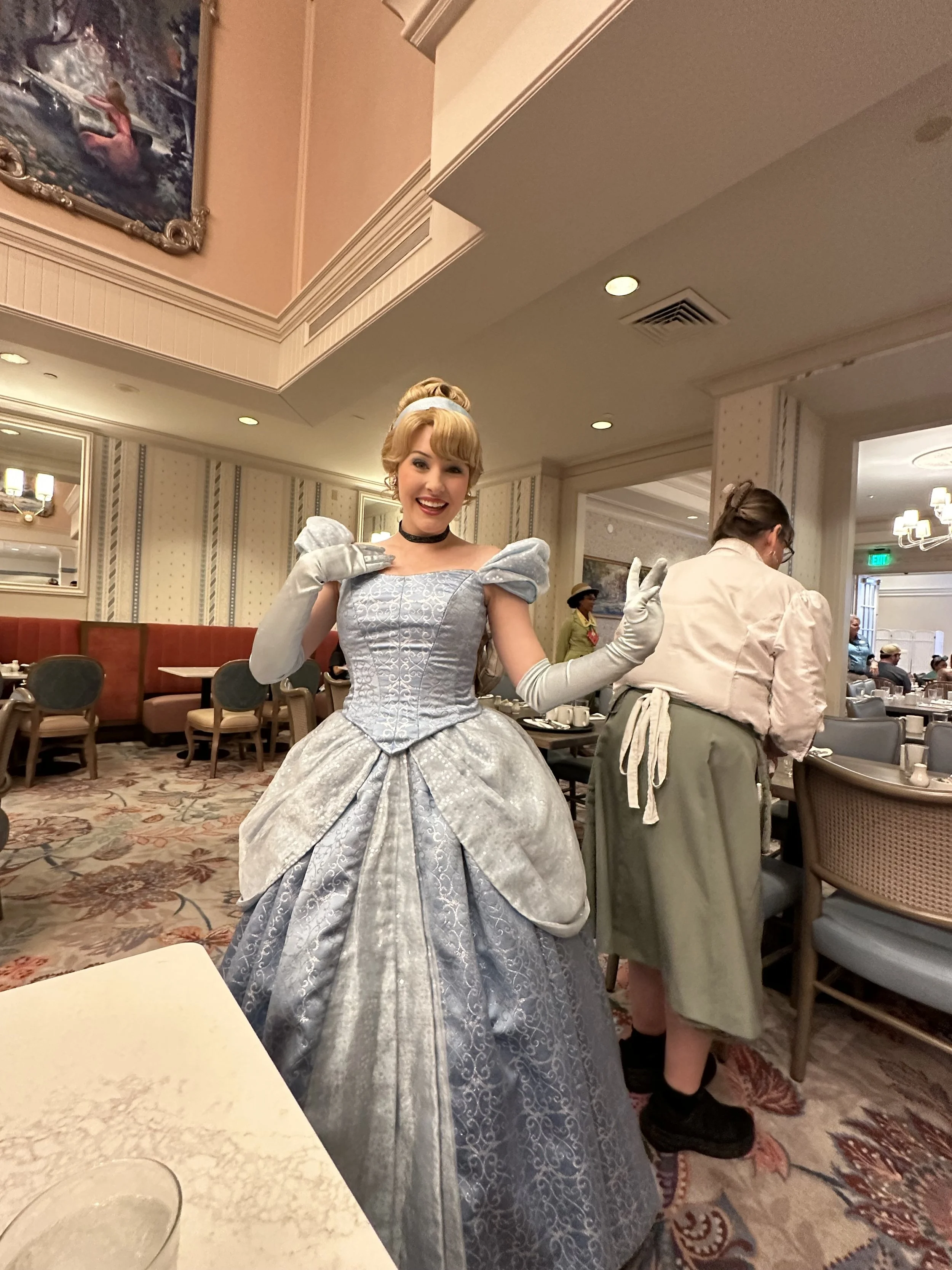 A woman dressed as a princess in a silver gown with puffed sleeves and gloves, smiling and giving a peace sign at a restaurant.