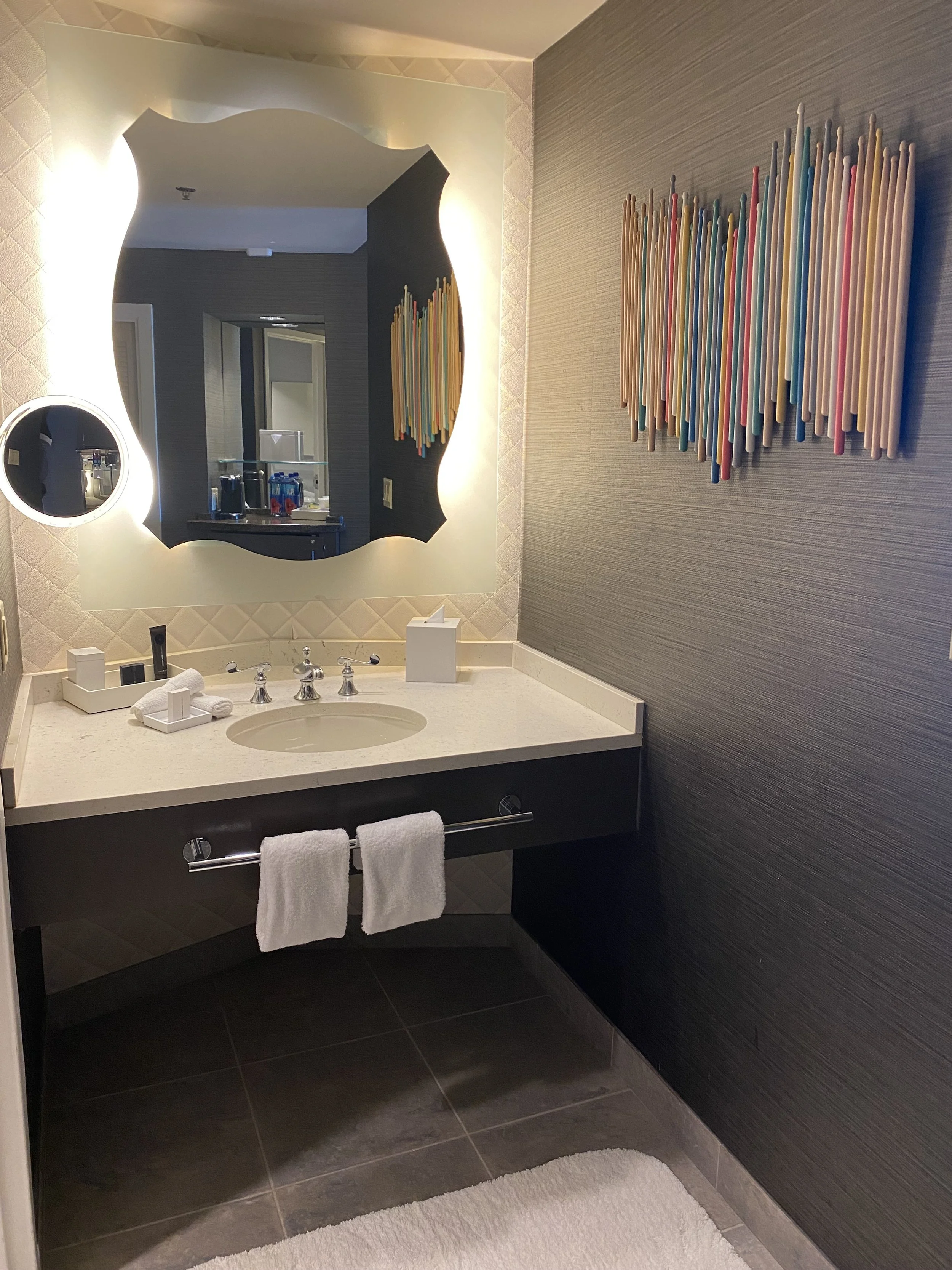 A modern bathroom with a backlit mirror, a smaller circular mirror, colorful artwork on the wall, a white countertop with a sink, and two white towels hanging on a towel bar.