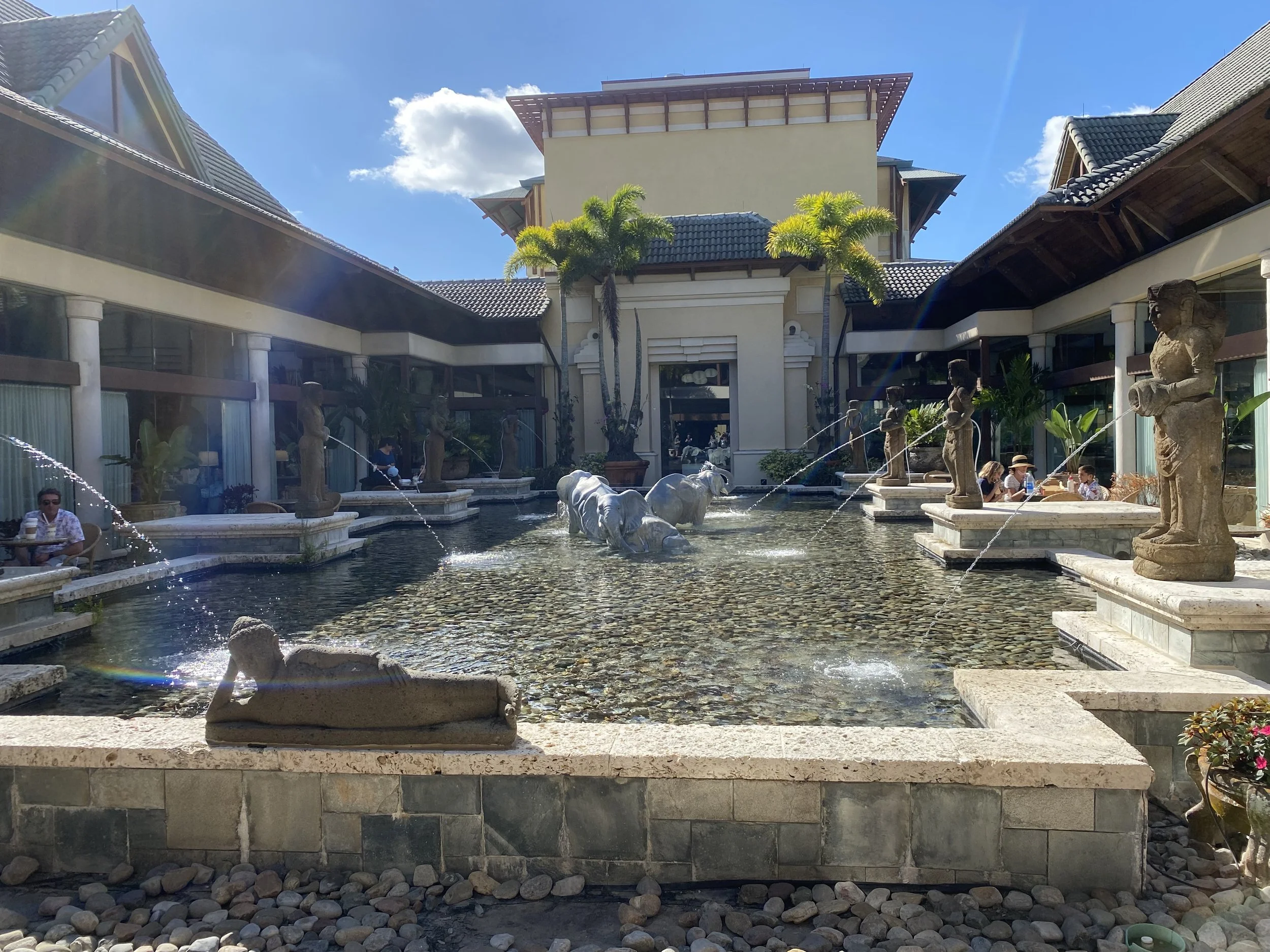 A decorative water fountain with stone statues of people and animals, surrounded by a building with large glass windows, palm trees, and people sitting nearby, under a clear blue sky.