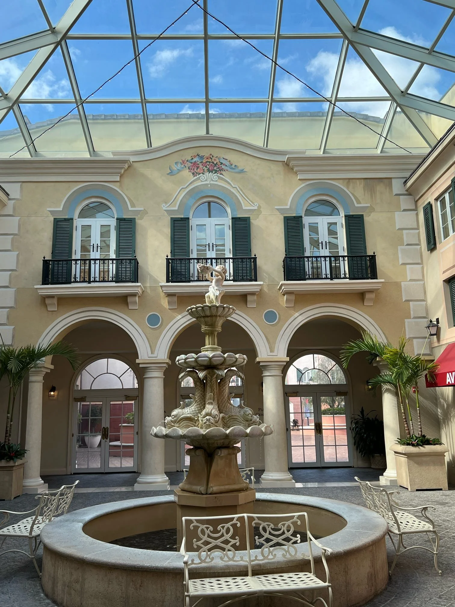 Indoor shopping mall with central fountain, two-story building with arched windows, balconies with green shutters, a glass ceiling, and potted plants.