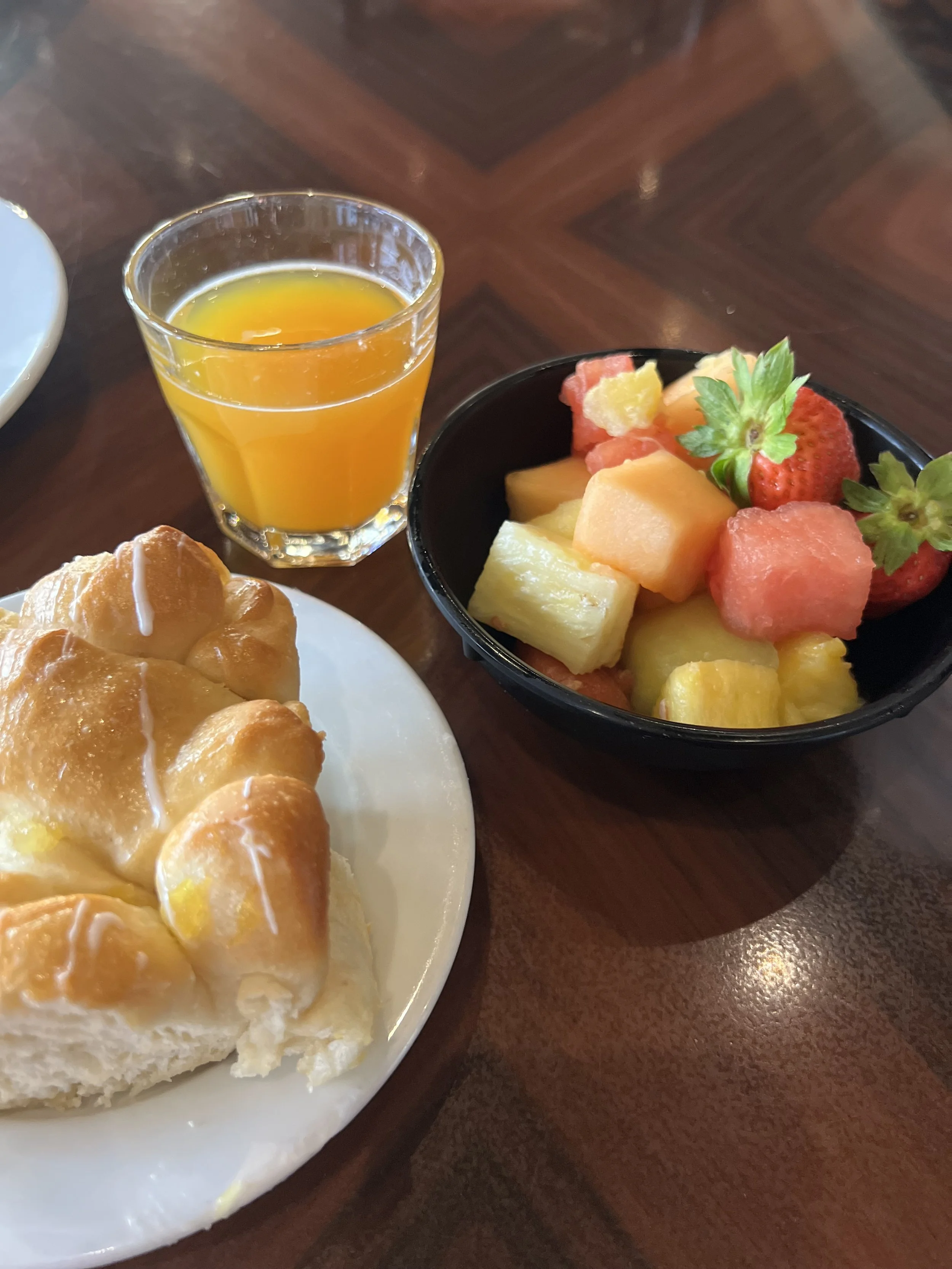 A breakfast spread featuring a glass of orange juice, a plate with a baked pain au chocolat, and a bowl of mixed fresh fruit including strawberries, melons, pineapple, and watermelon on a wooden table.